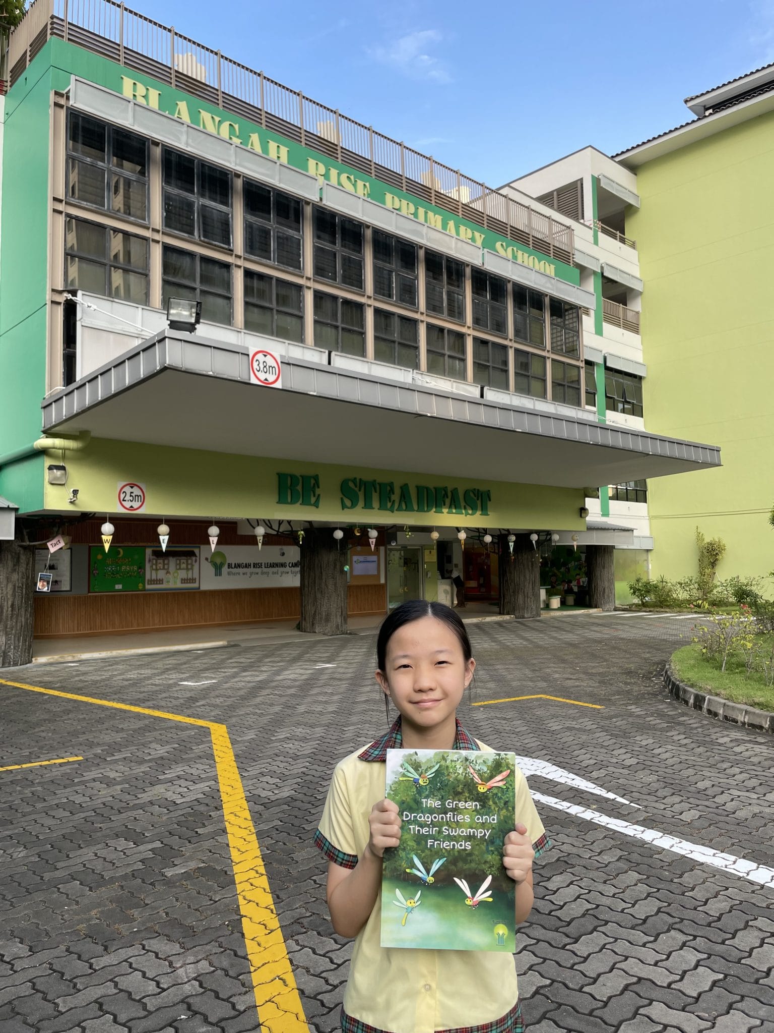 A girl holds a book titled "The Green Dragonflies and Their Swampy Friends" in front of Blangah Rise Primary School.