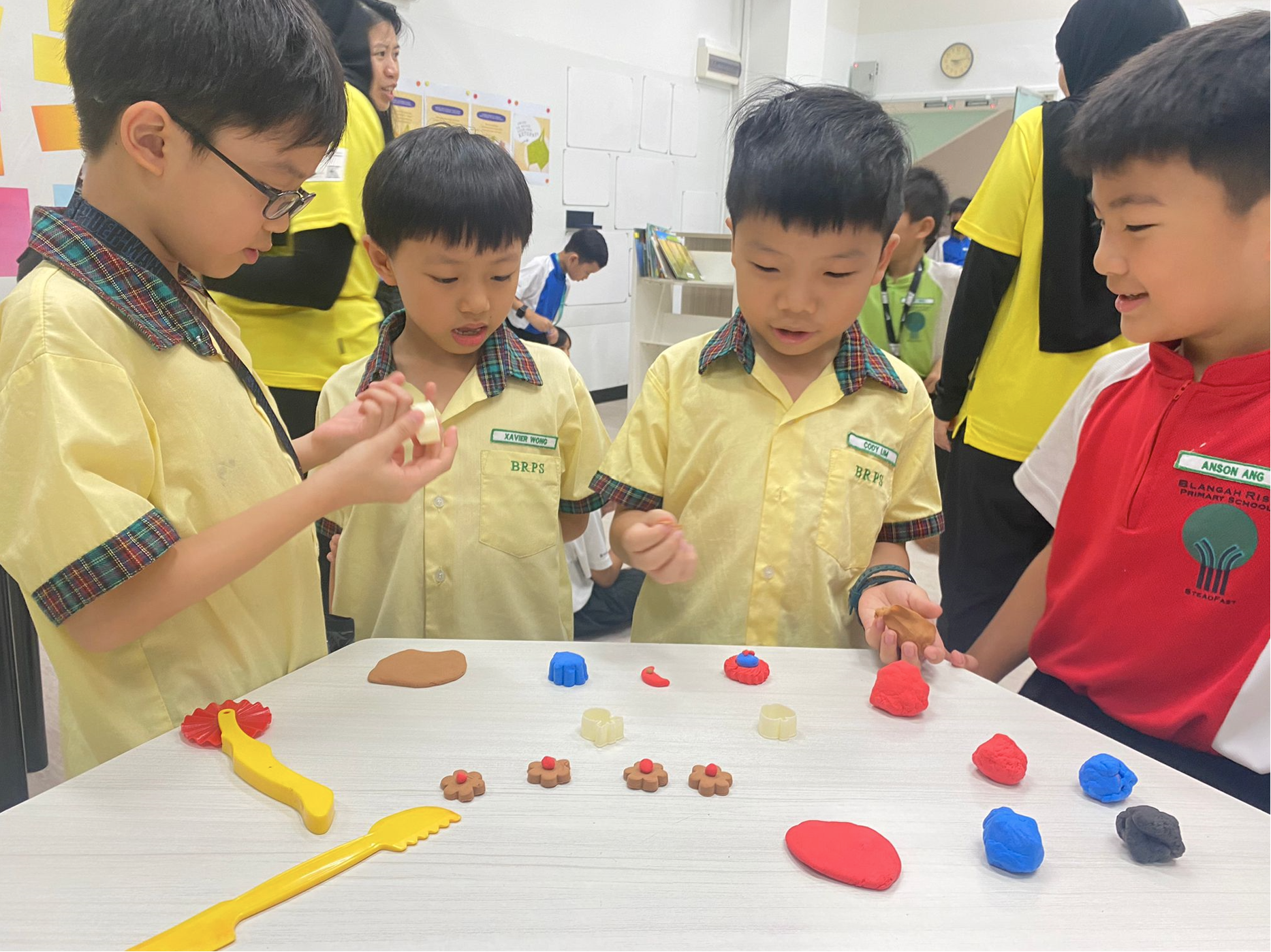 Four young school boys in uniforms gather around a table covered with colorful clay sculptures they are molding.