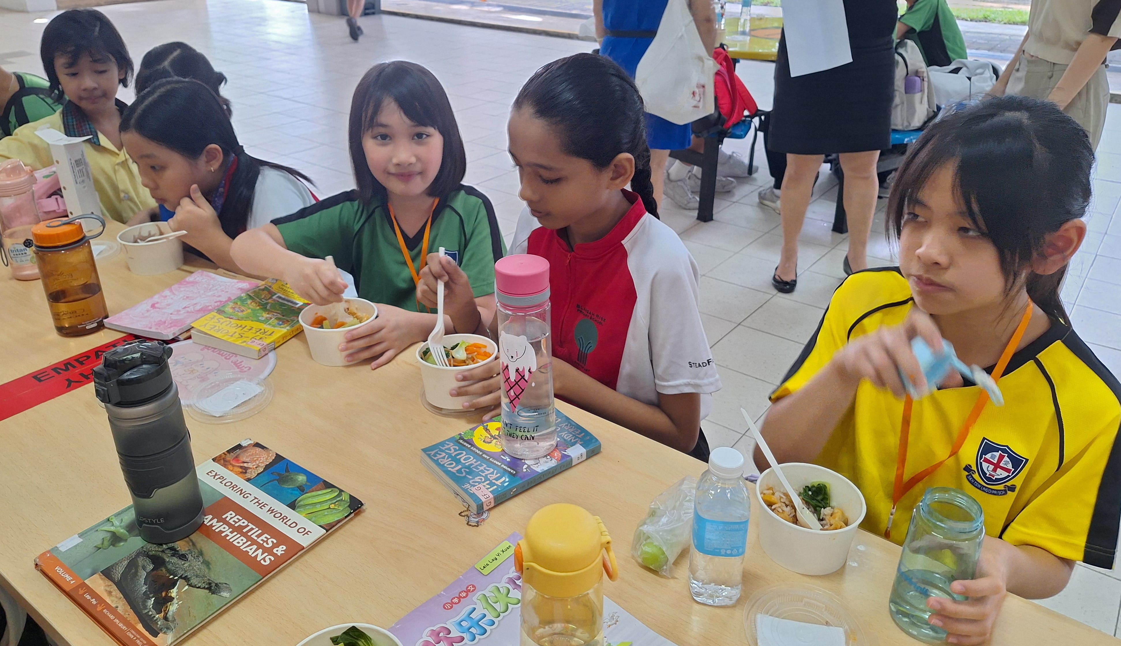 Several school children in uniforms eat meals at a table covered with books, water bottles, and food bowls.