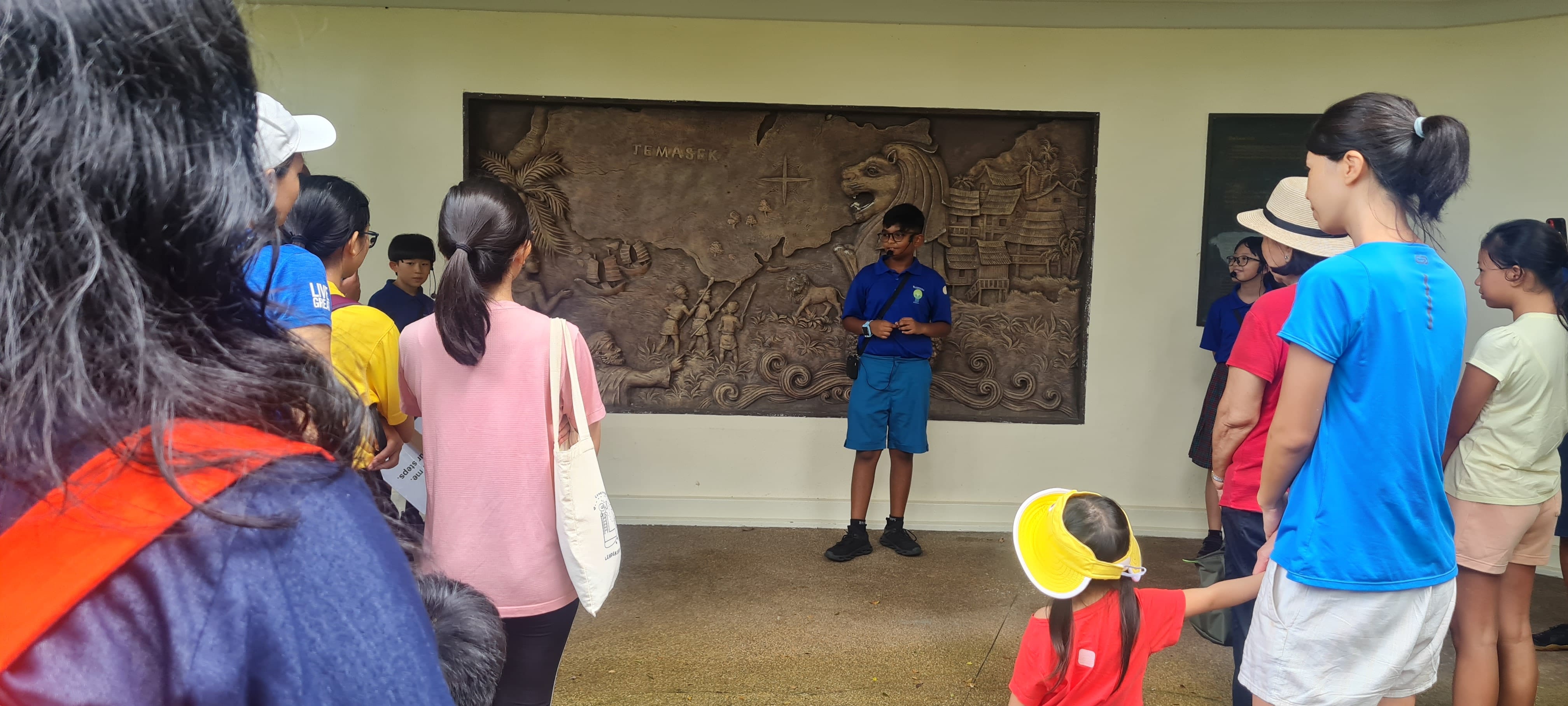 A guide in blue stands before a bronze Temasek relief mural, presenting to a group of adults and children.