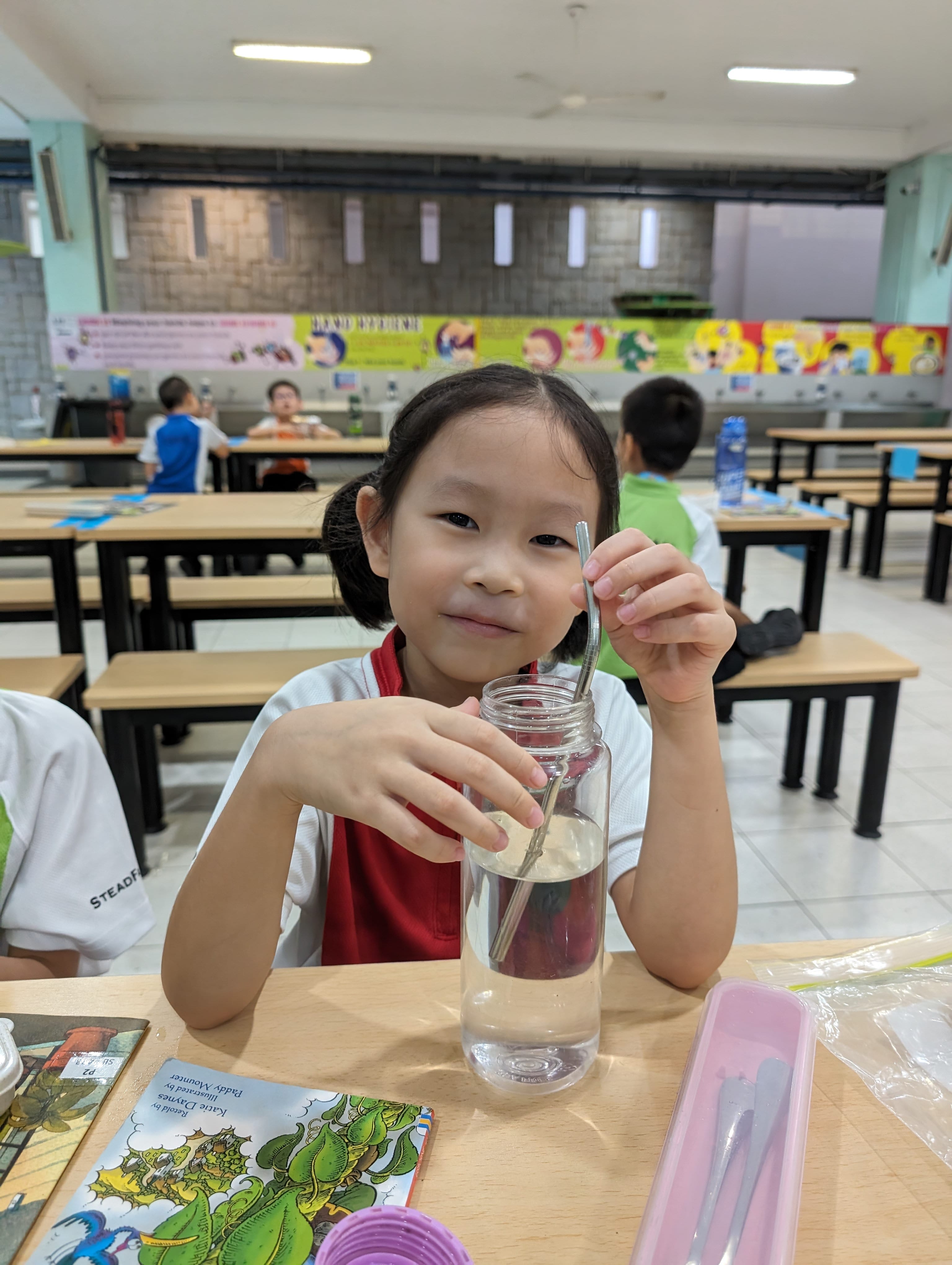 Young girl smiling at a school cafeteria table, holding a metal straw in a clear glass water bottle.