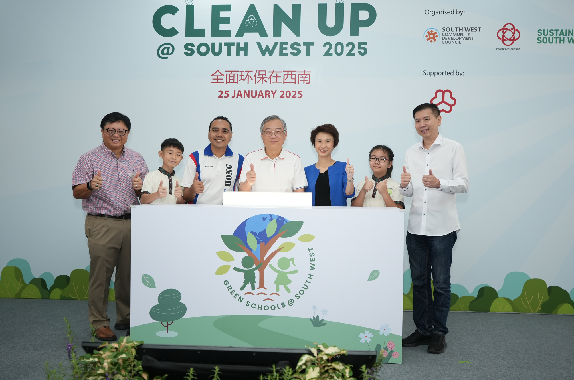 Seven people pose giving thumbs up behind a Green Schools banner under Clean Up @ South West 2025 signage.