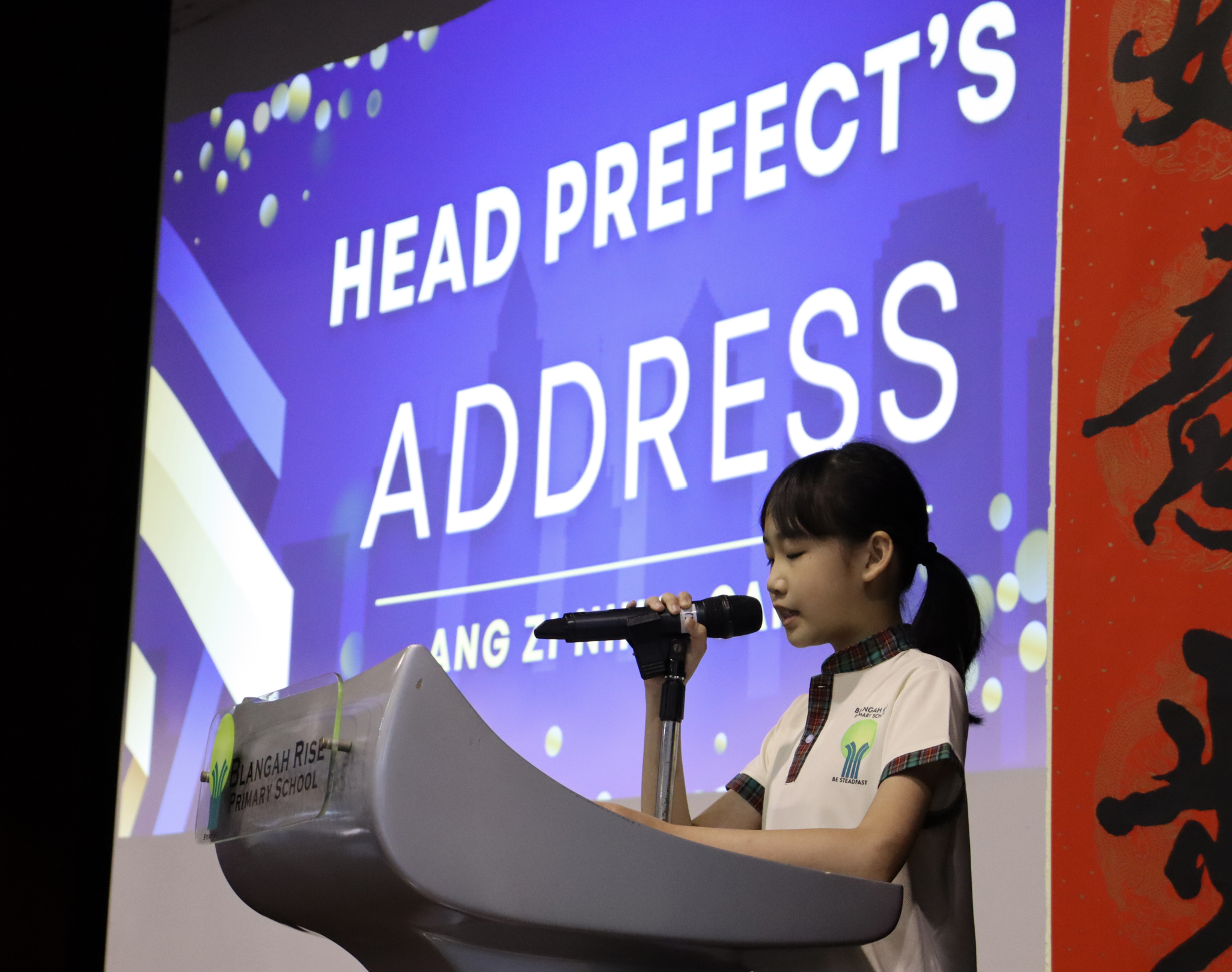 Student speaks into a microphone at a podium with a large screen behind reading HEAD PREFECT'S ADDRESS