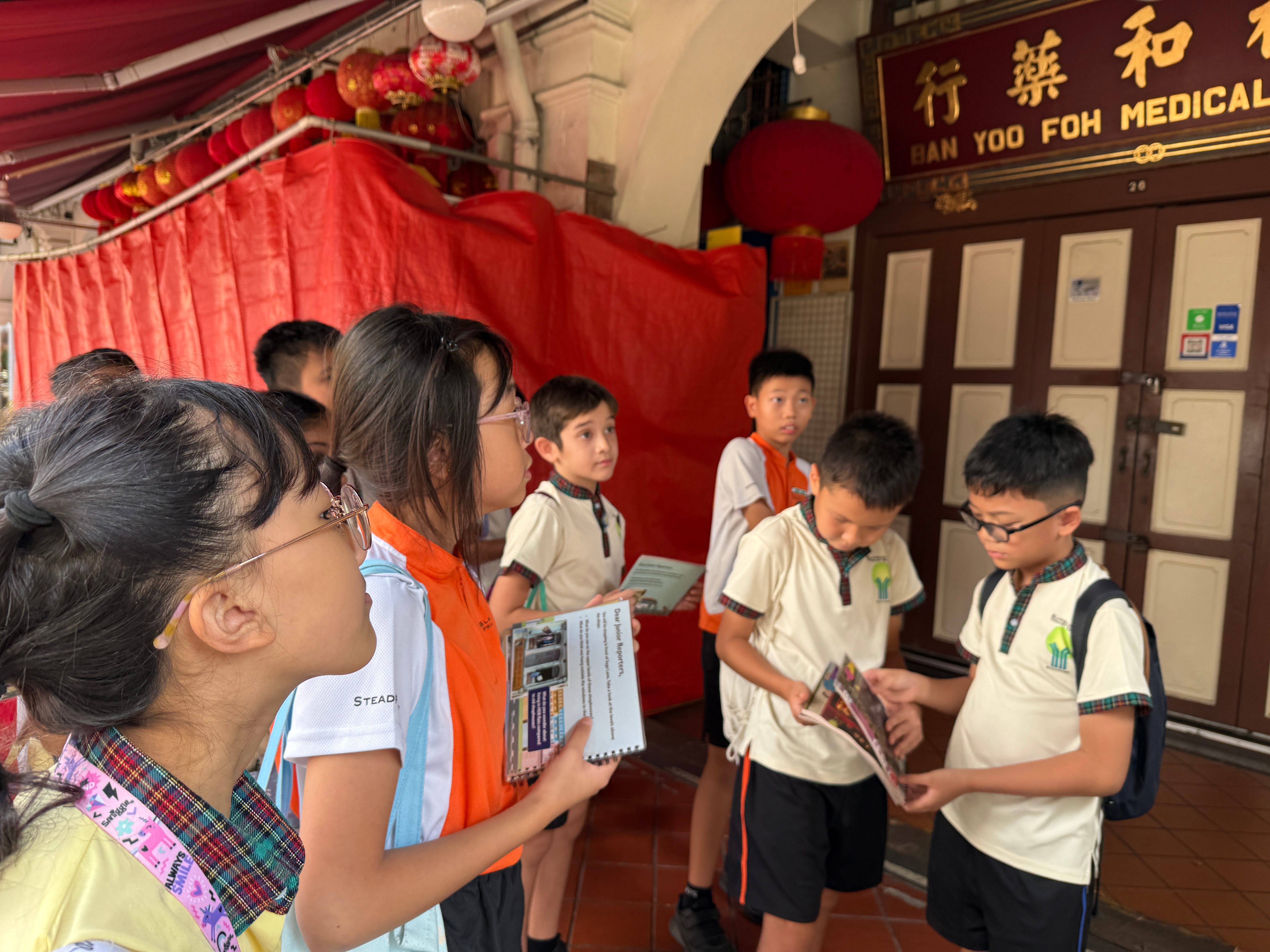 Group of schoolchildren holding pamphlets outside a doorway with red lanterns and a Chinese sign above.