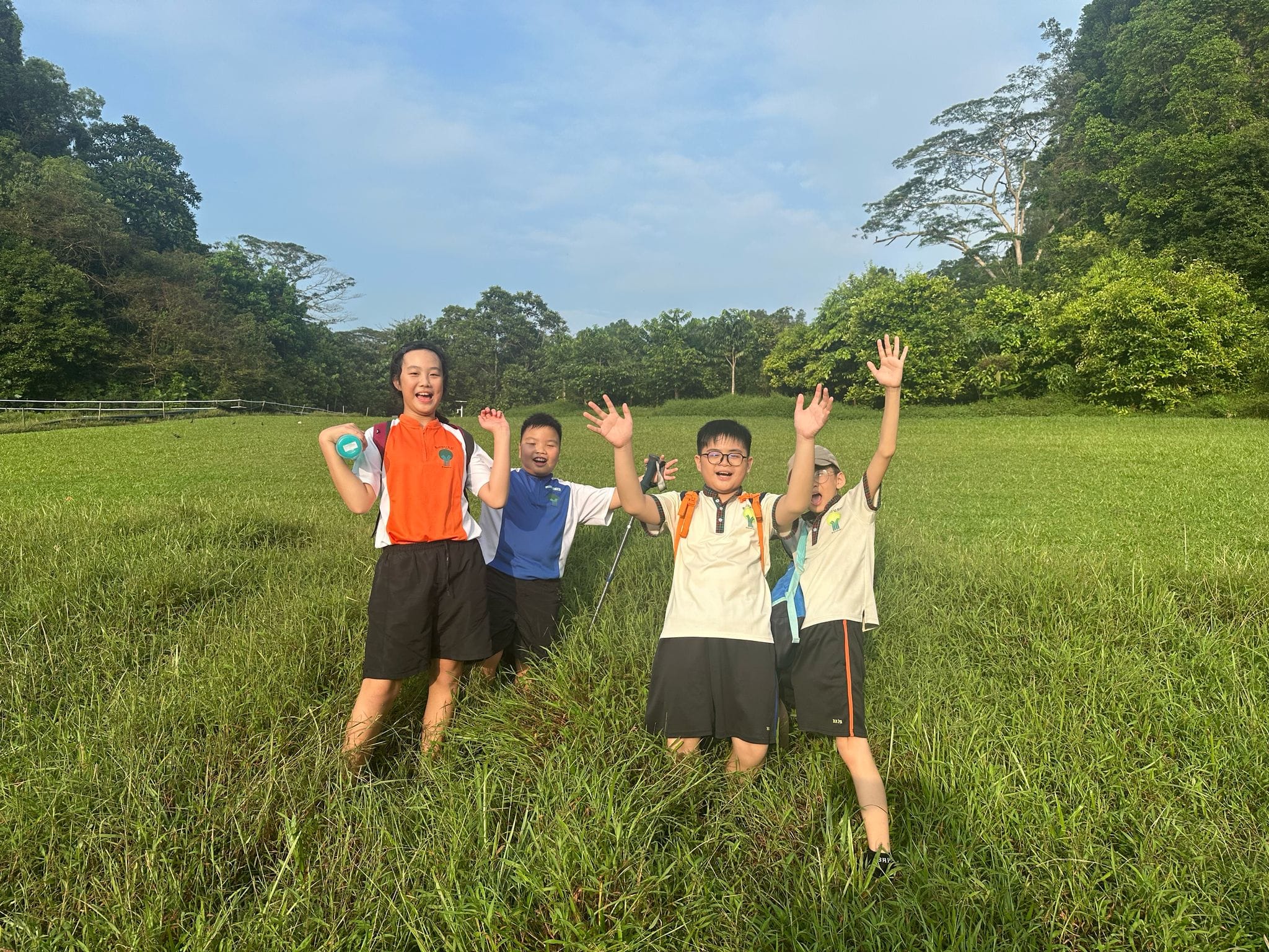 Four cheerful children pose with raised arms in a grassy field surrounded by green trees under a blue sky.