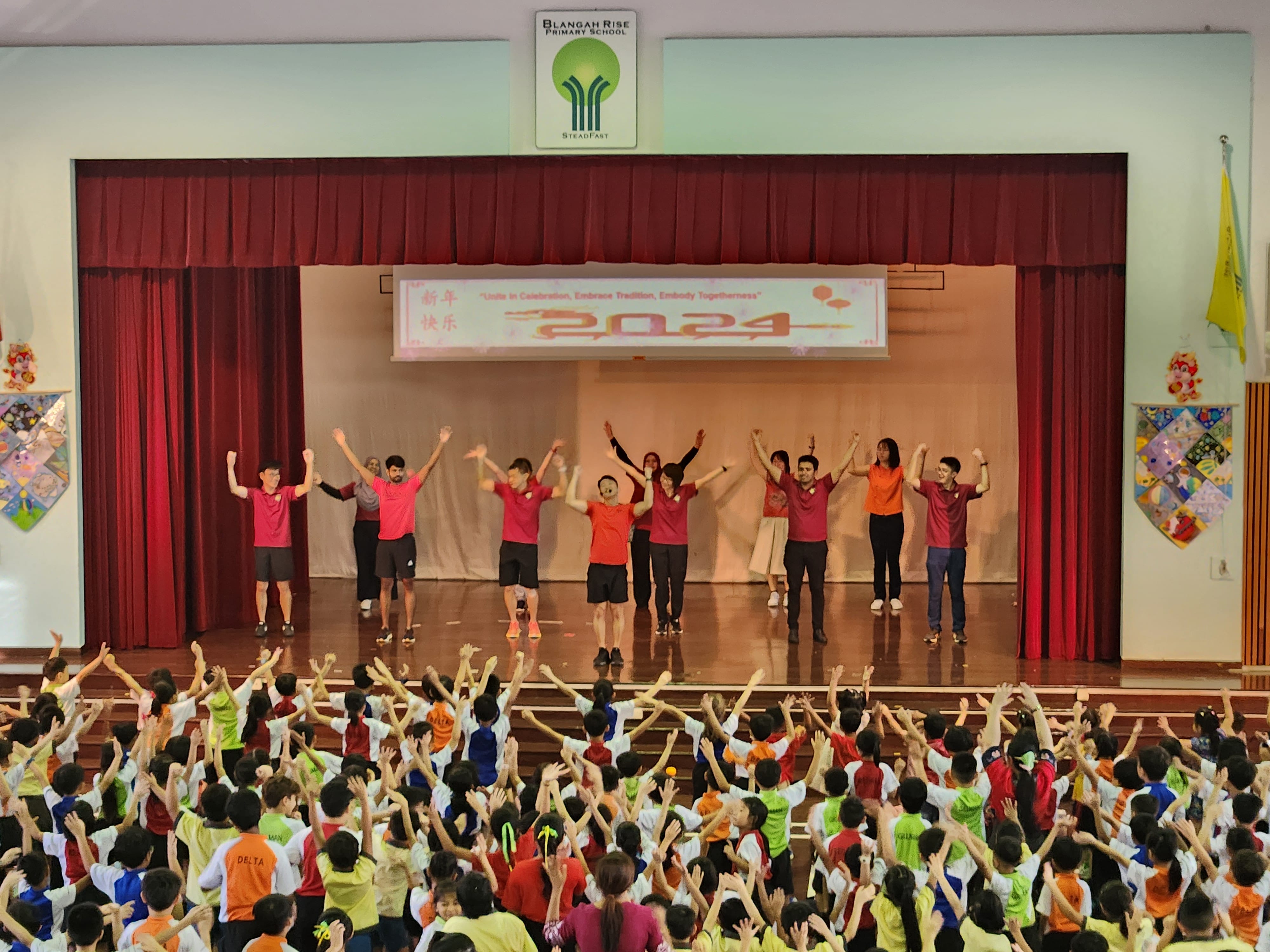 Teachers on stage raise hands before a crowd of students at a 2024 Lunar New Year school celebration.