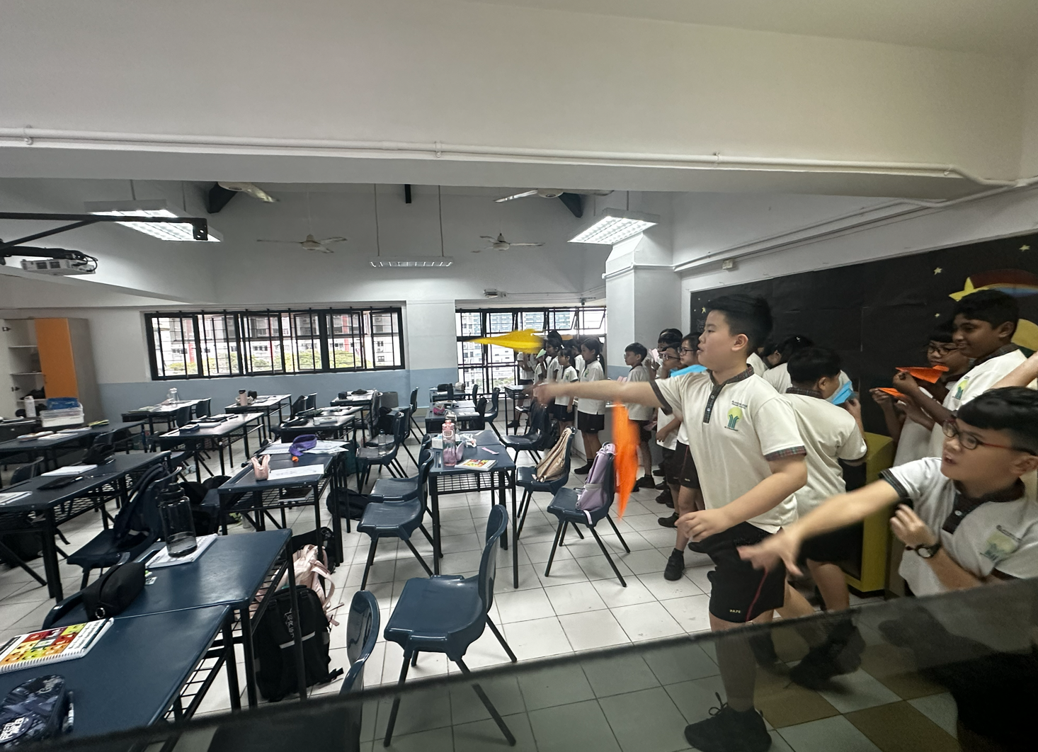 Students in school uniforms throw colorful paper planes across a classroom with blue desks and tiled floors.