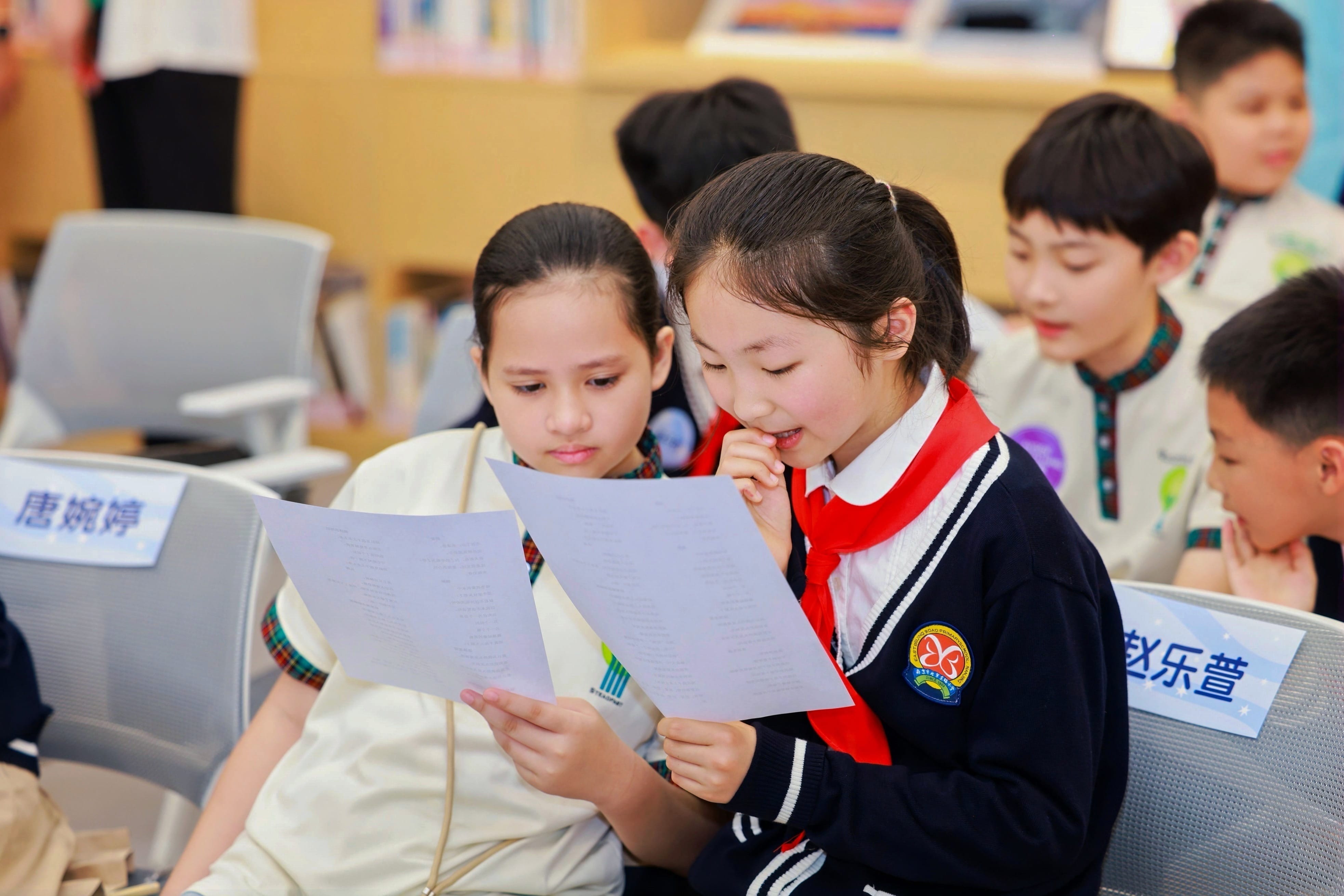 Two young girls in school uniforms read printed papers together, seated among other children with Chinese name placards.
