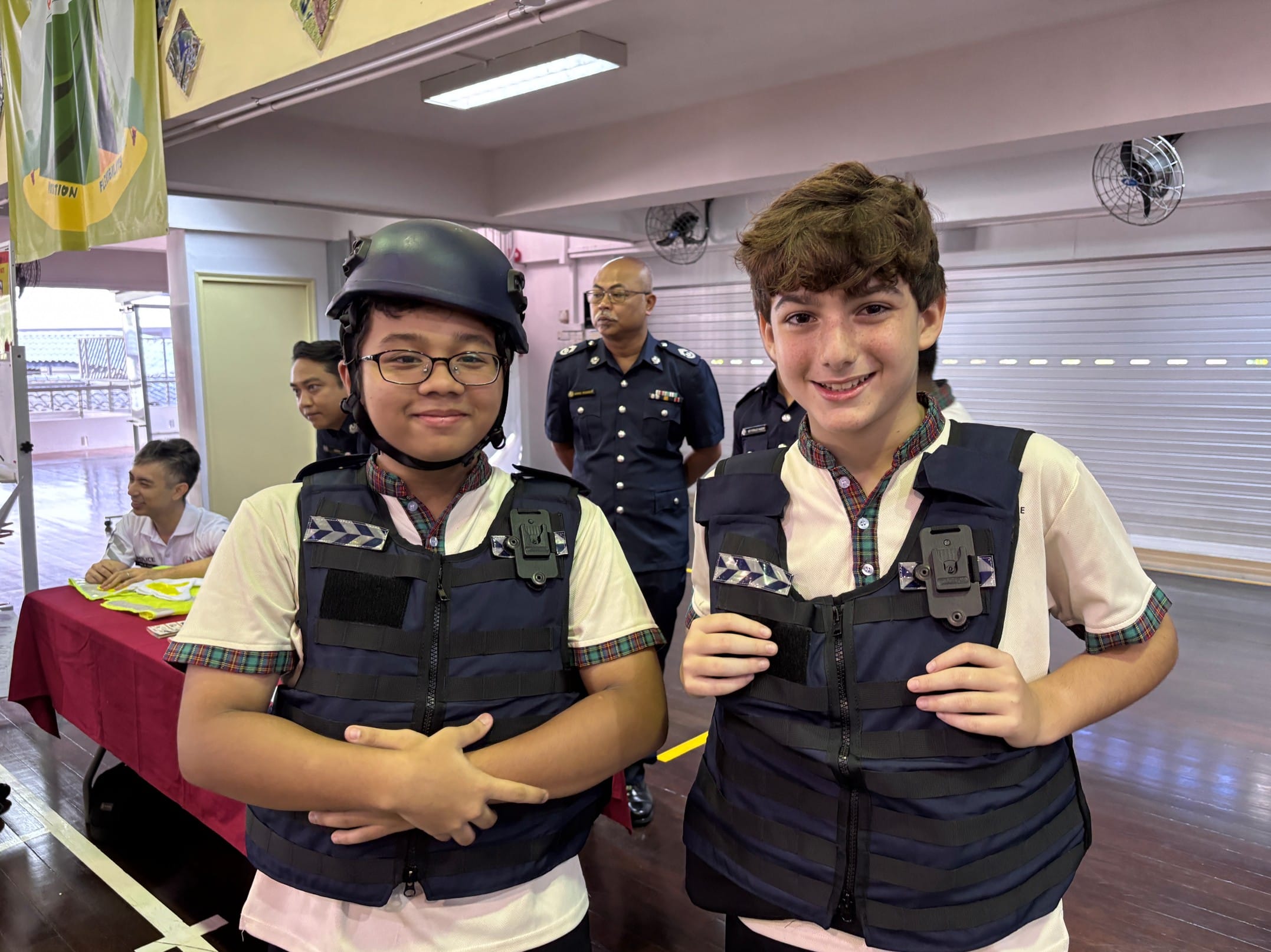 Two smiling children wearing tactical vests, one with a helmet, stand indoors with uniformed officers in the background.