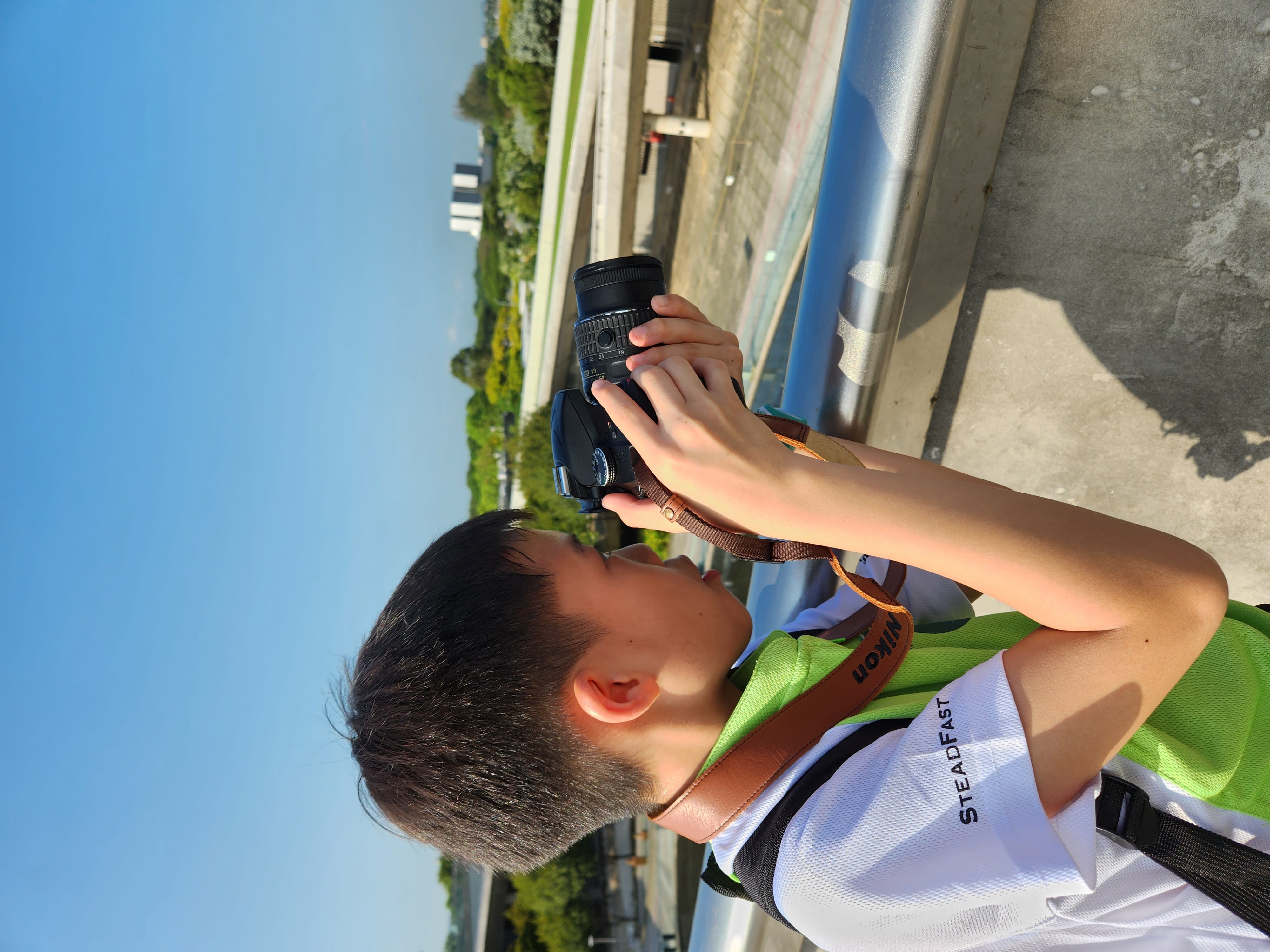 A young boy in a green vest holds a Nikon DSLR camera up to shoot outdoors on a rooftop under blue sky.