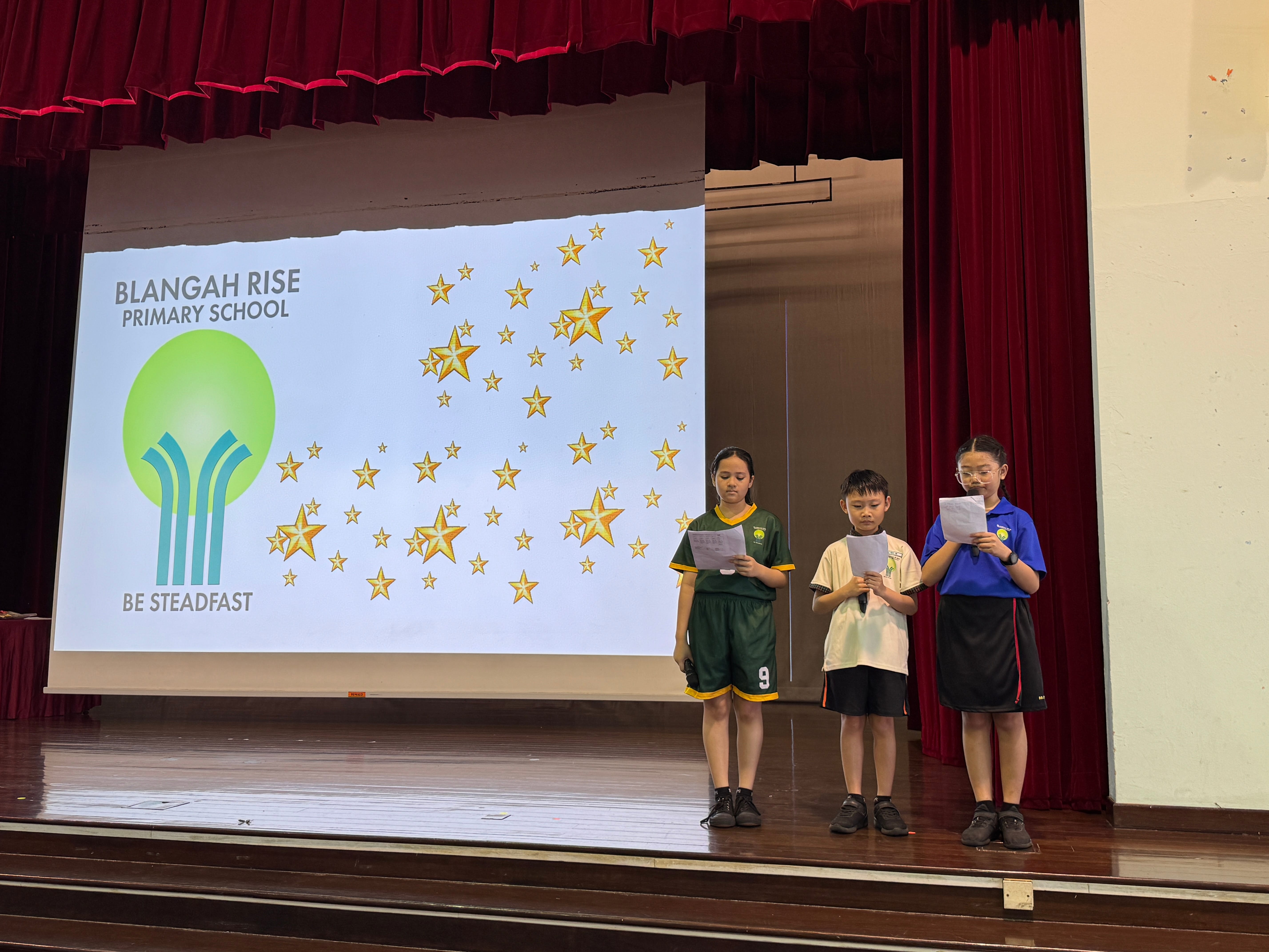 Three schoolchildren on stage reading papers beside a screen showing Blangah Rise Primary School and stars.