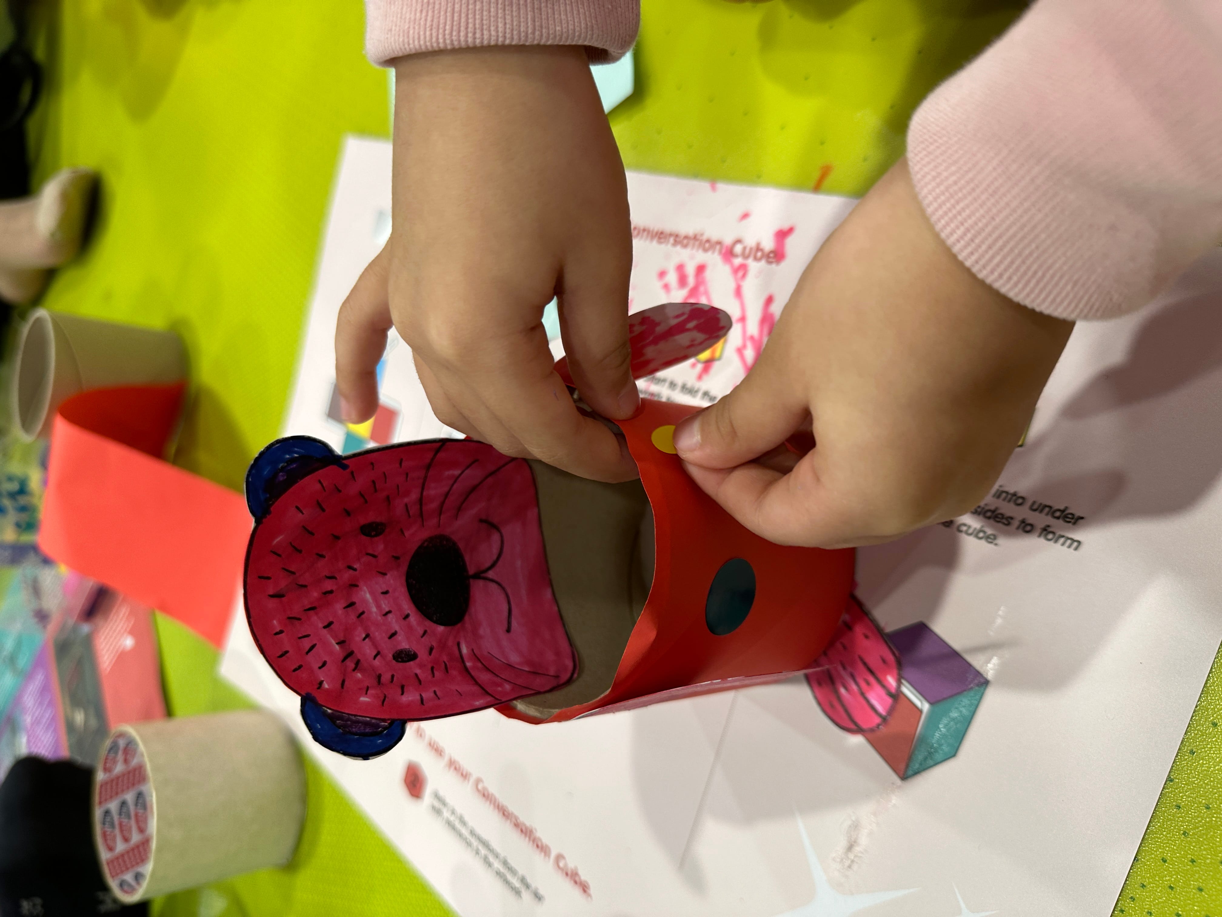 Child’s hands assemble a paper bear craft with a red body on a green table, with tape and supplies nearby.