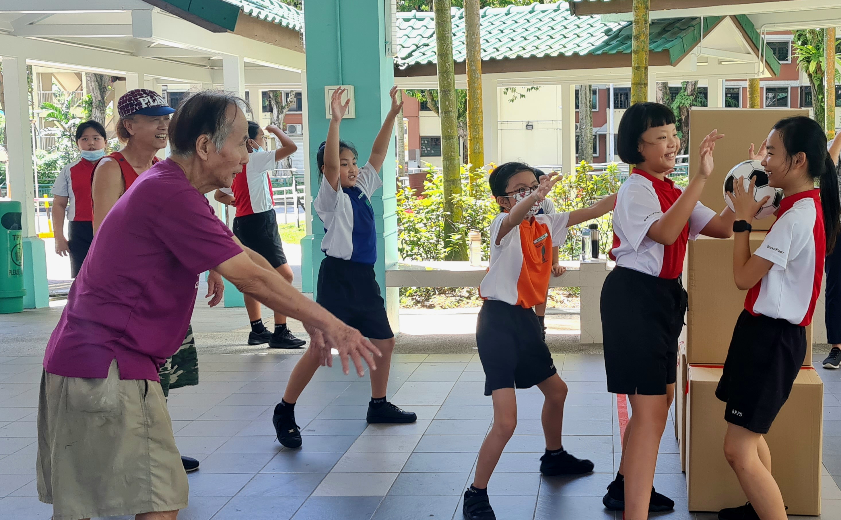 Elderly man and school children dancing together outdoors under a covered walkway, smiling and raising their arms.