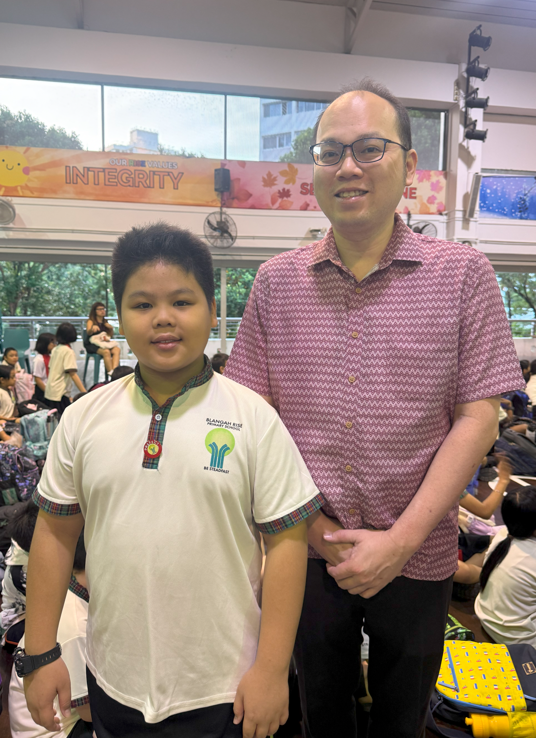 A young student in a Blangah Rise Primary School uniform stands beside an adult man in a pink shirt inside a school hall.