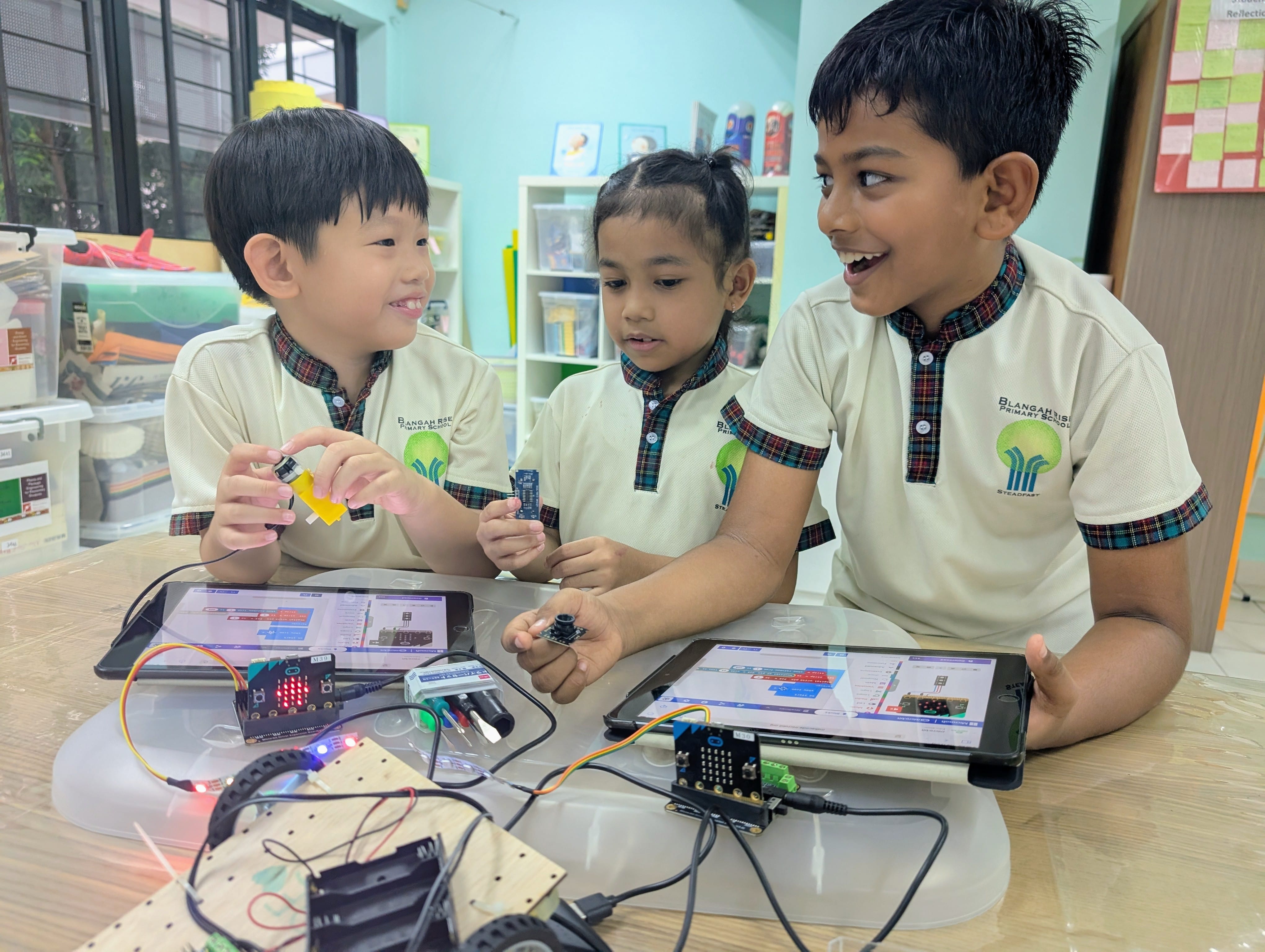 Three primary school children in uniforms work together on electronics and coding using tablets and circuit boards.