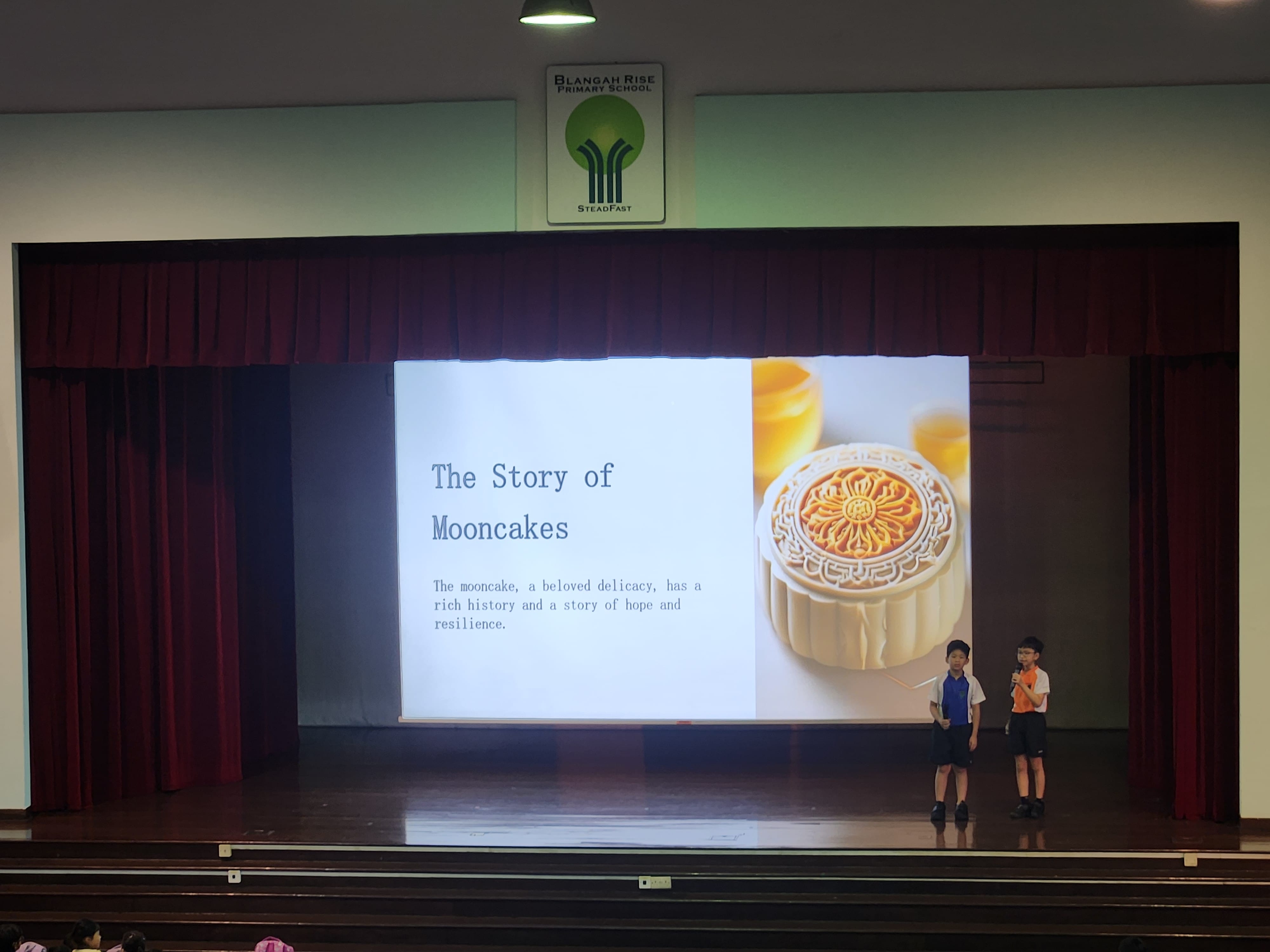 Two children stand on a stage beside a projected slide titled The Story of Mooncakes with a mooncake photo.