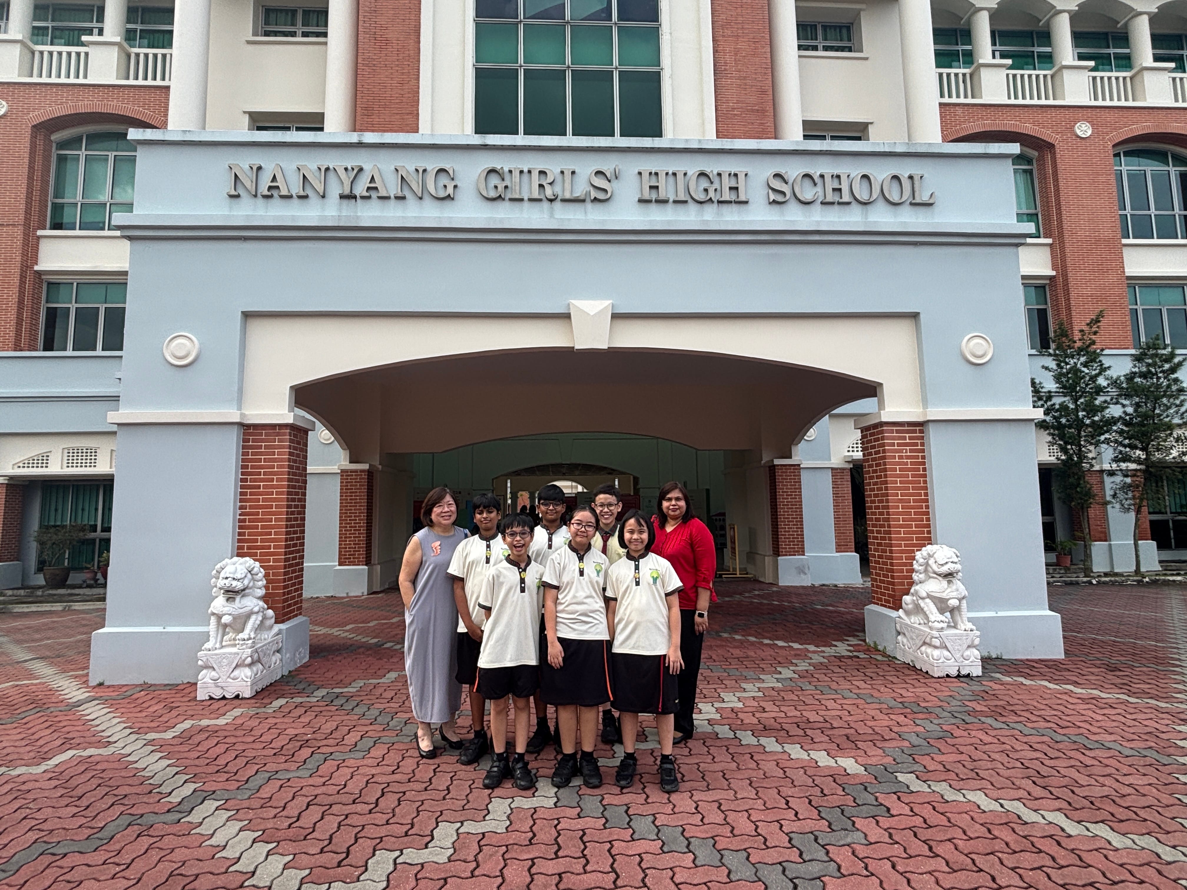 A group of students and two adults pose in front of Nanyang Girls' High School entrance with stone lion statues.