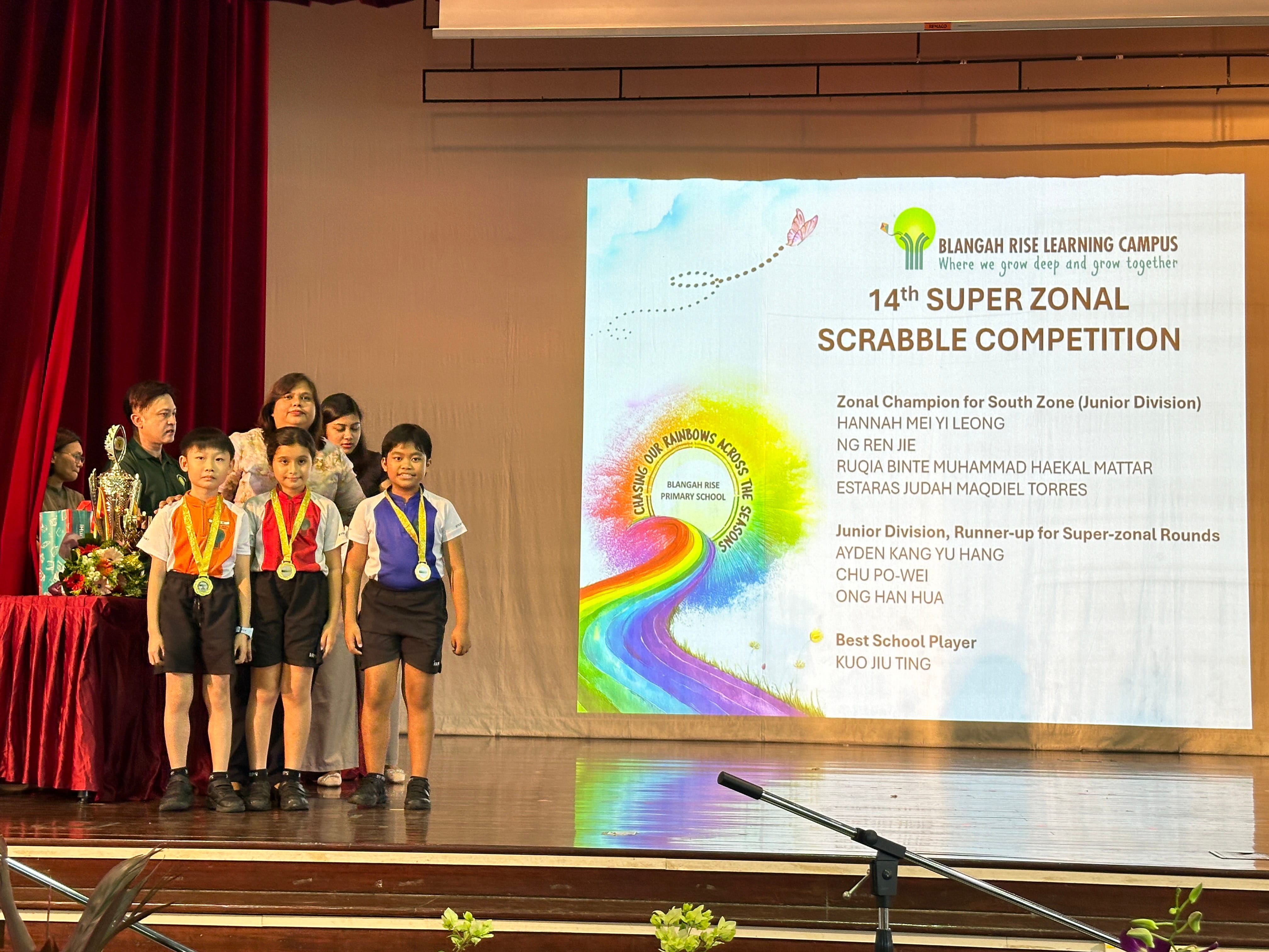 Three medal-wearing children stand on a stage beside adults next to a 14th Super Zonal Scrabble Competition presentation slide.