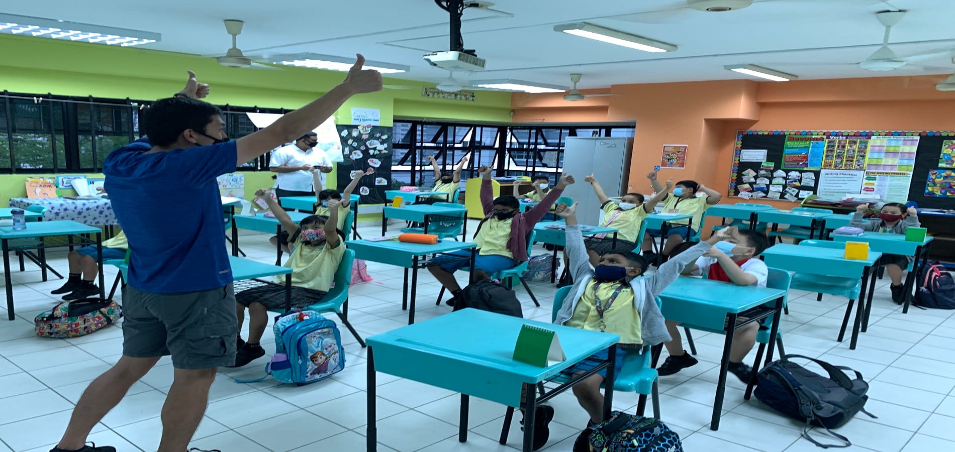 A masked teacher gives a thumbs up to students wearing masks who raise their hands in a bright classroom.
