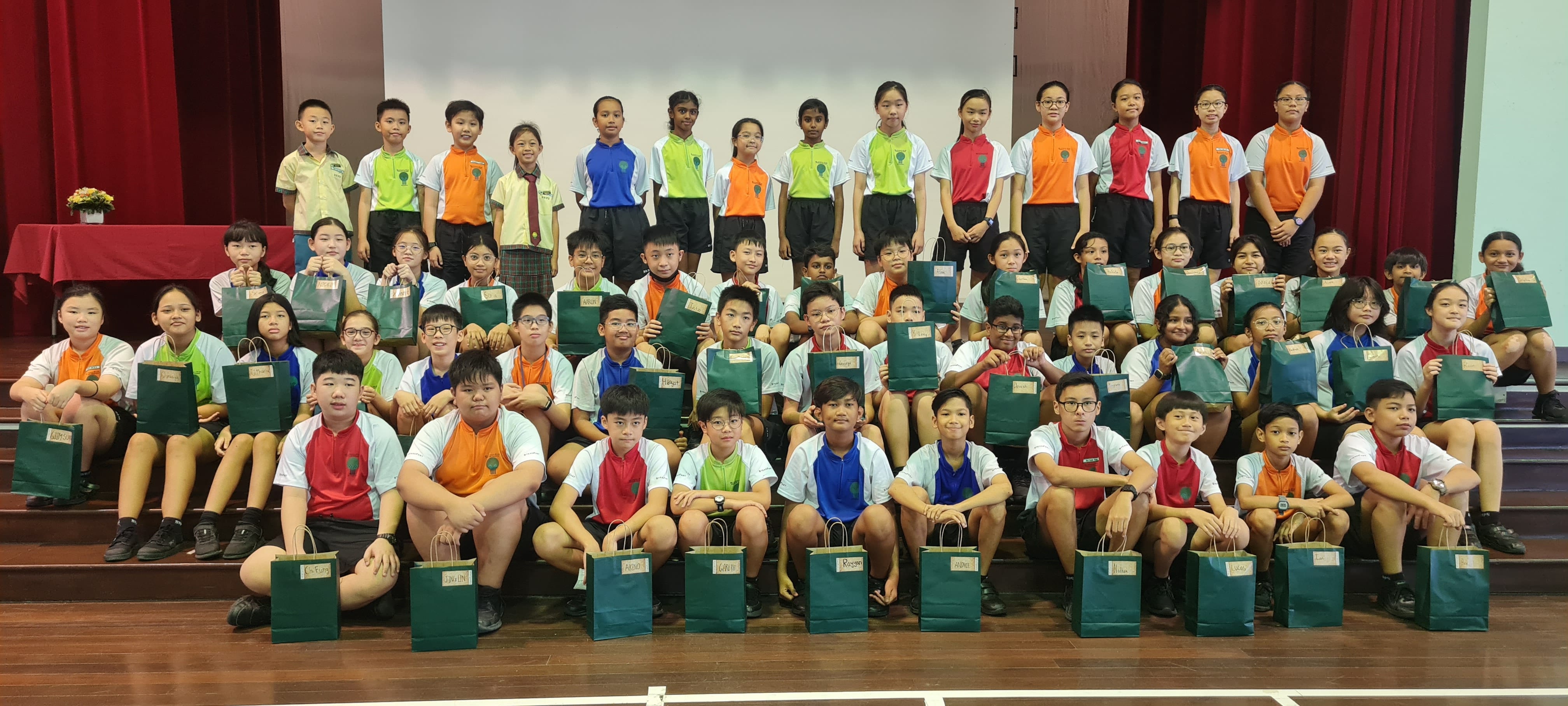Large group of school children in colorful uniforms posing on a stage, many holding green gift bags.