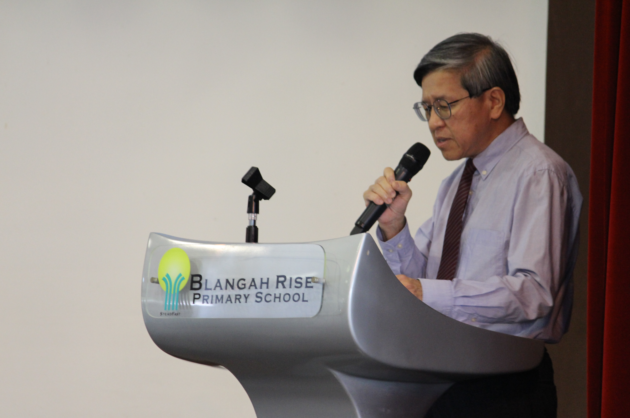 Man in glasses and tie speaking into a microphone at a Blangah Rise Primary School podium.