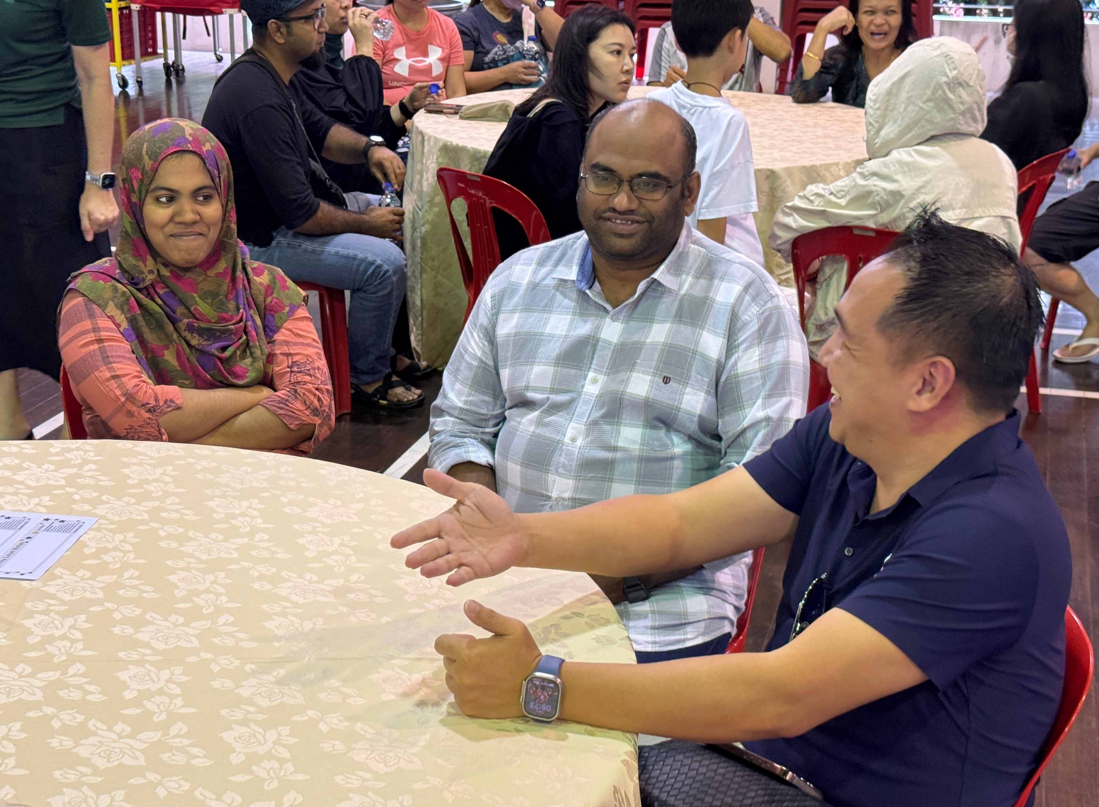 Three people sit at a round table talking, including a woman in a hijab and a man gesturing with his hand.