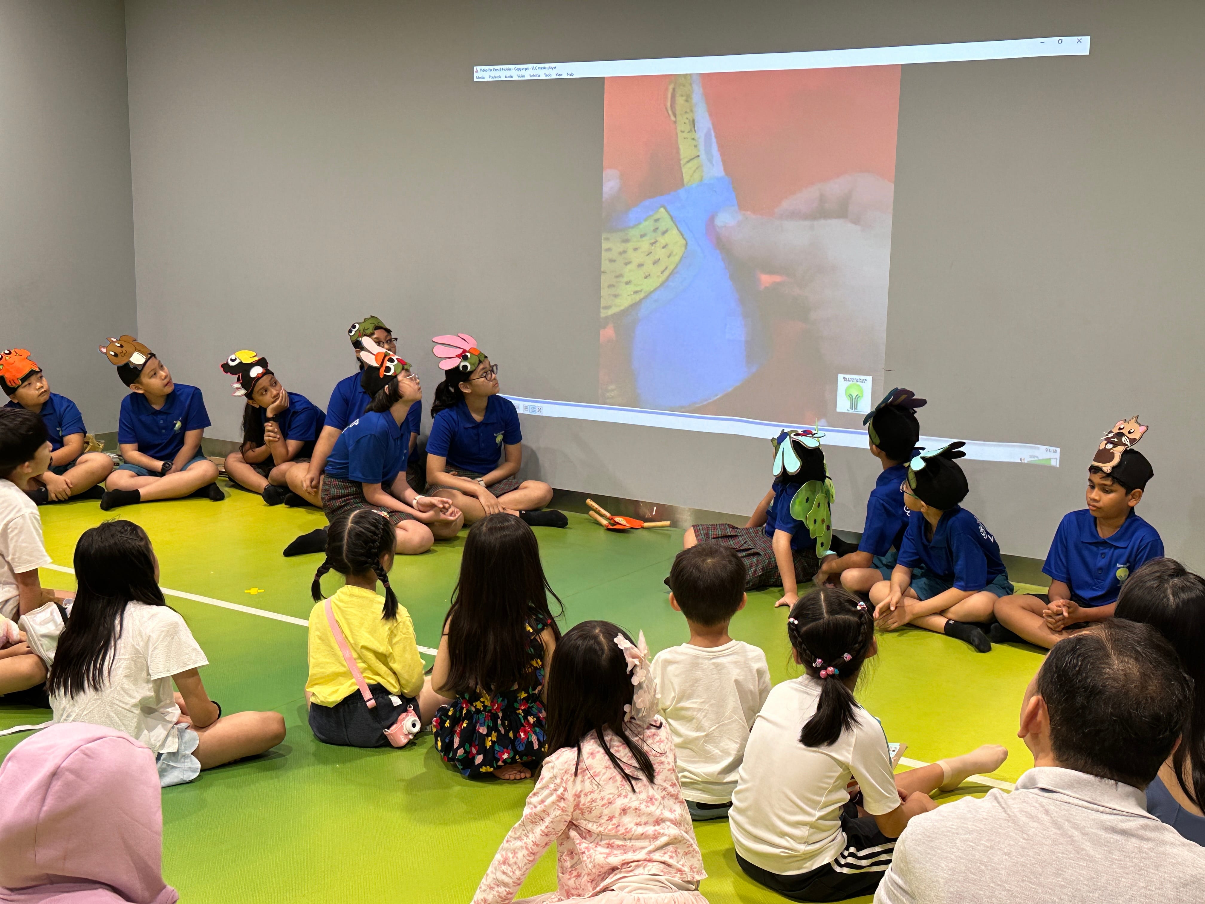 Children sitting on green floor watch a wall projection while some wear animal-themed headbands.