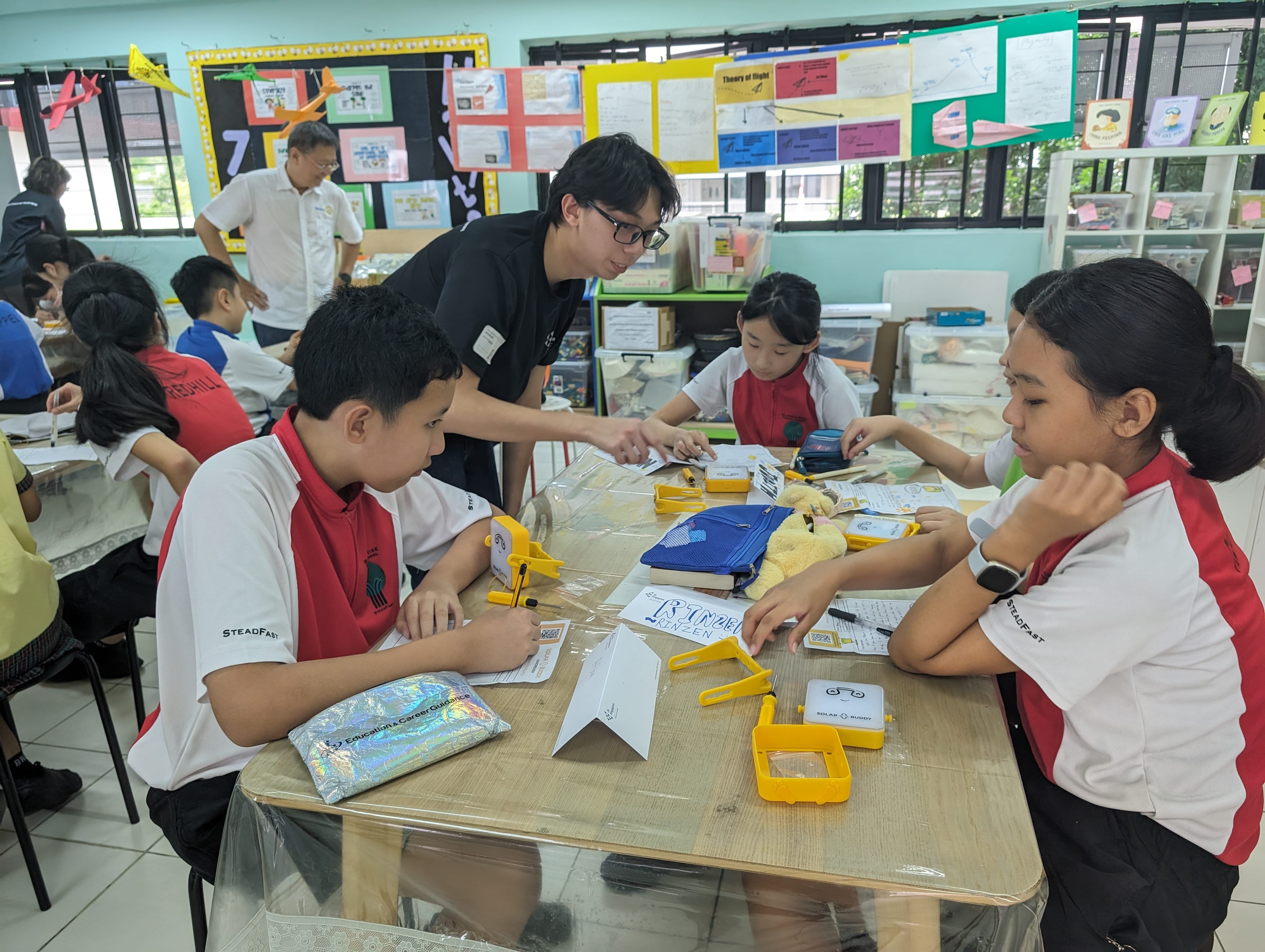 Students in red and white uniforms work at a table with yellow tools while a teacher in black leans over to assist.