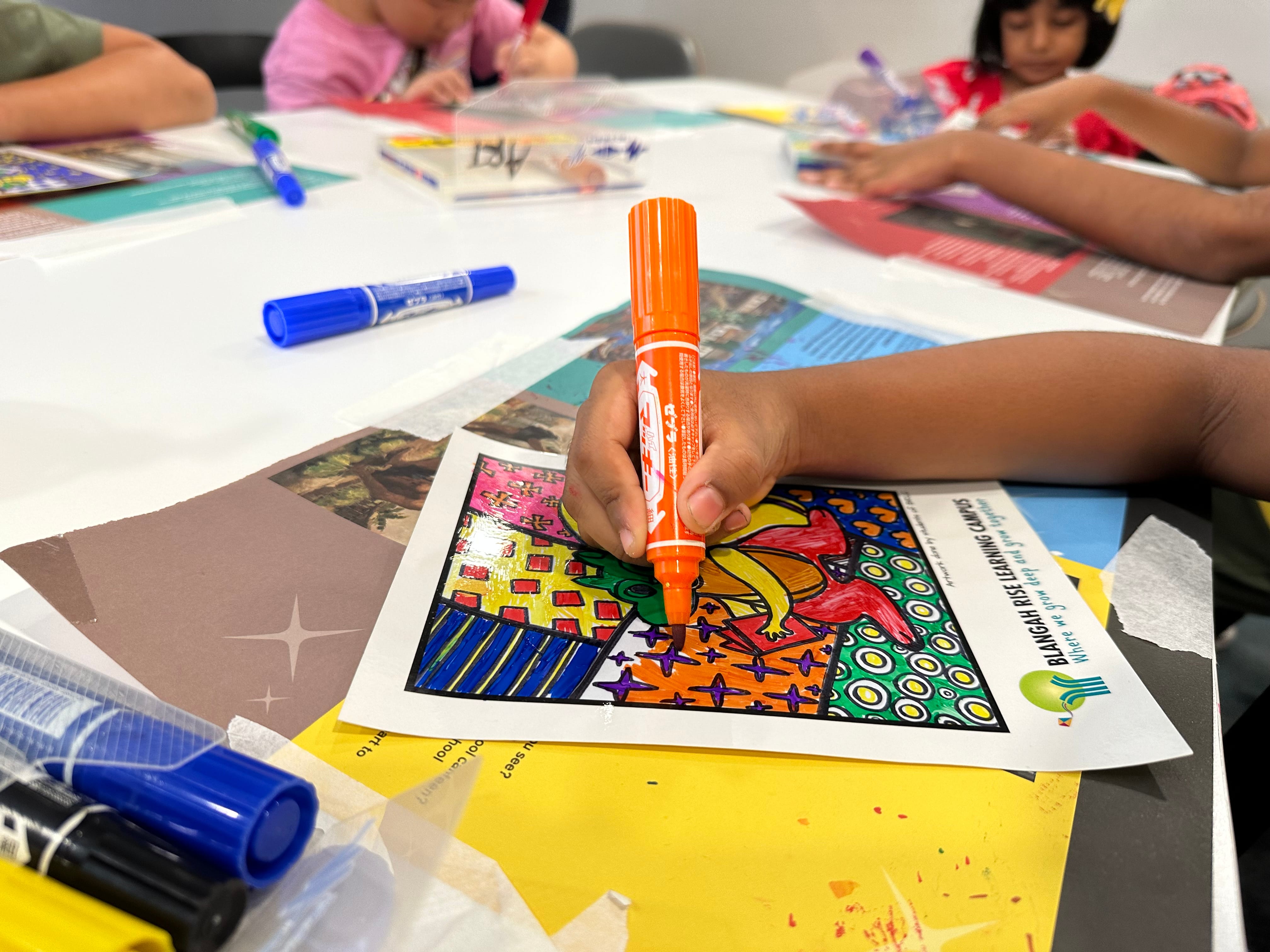 A child's hand holds an orange marker coloring a vibrant printed artwork at a table with other children and markers.