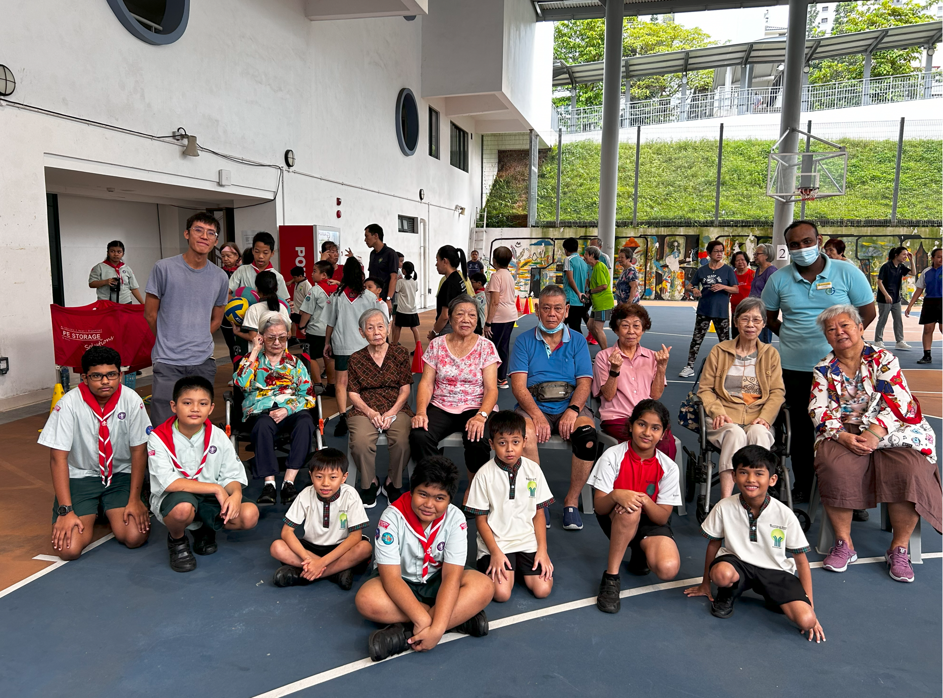 Group photo of elderly people seated with schoolchildren kneeling in front on a covered outdoor court.