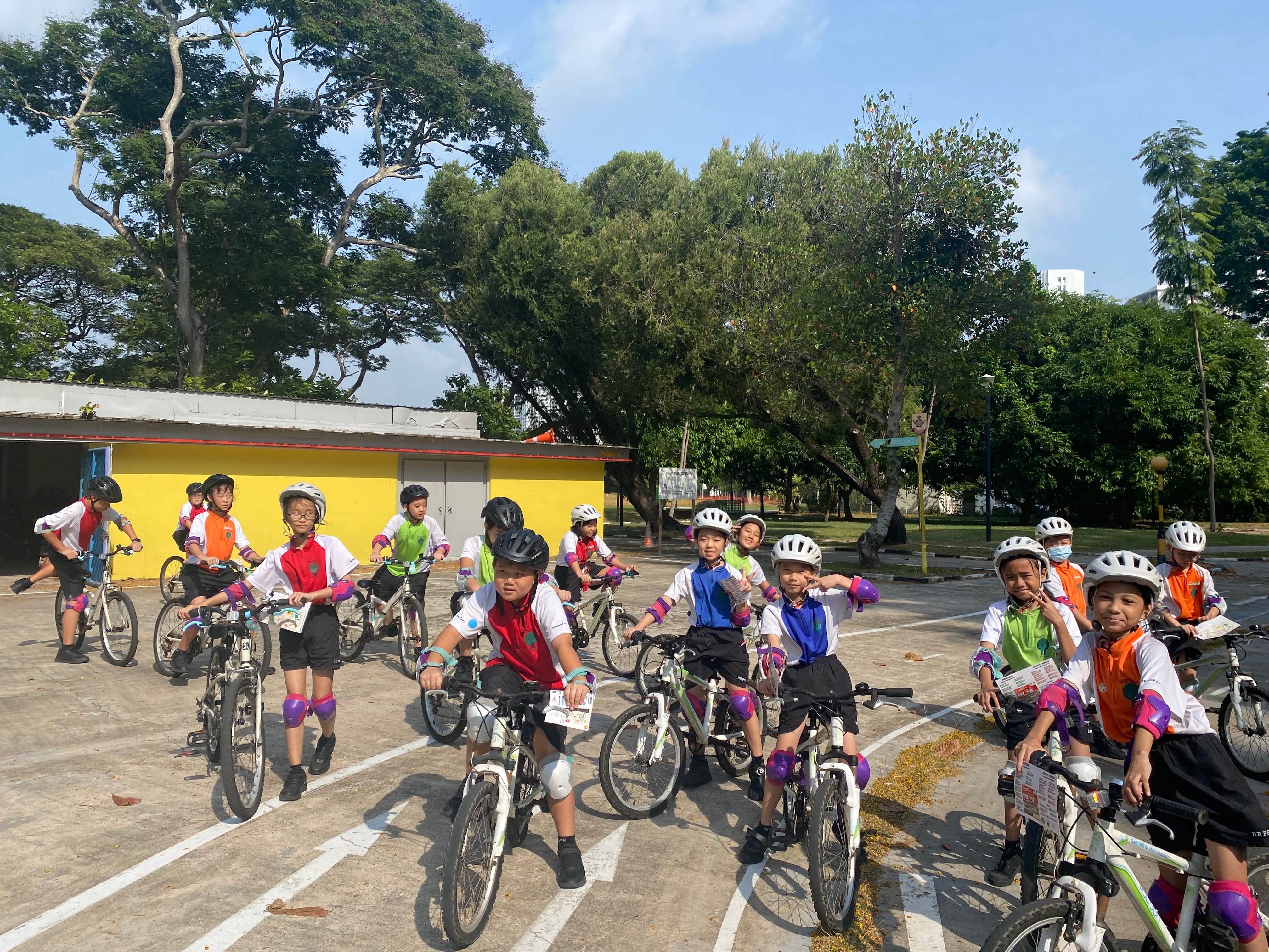 A group of young children wearing helmets and knee pads sit on bicycles in an outdoor paved area with trees.