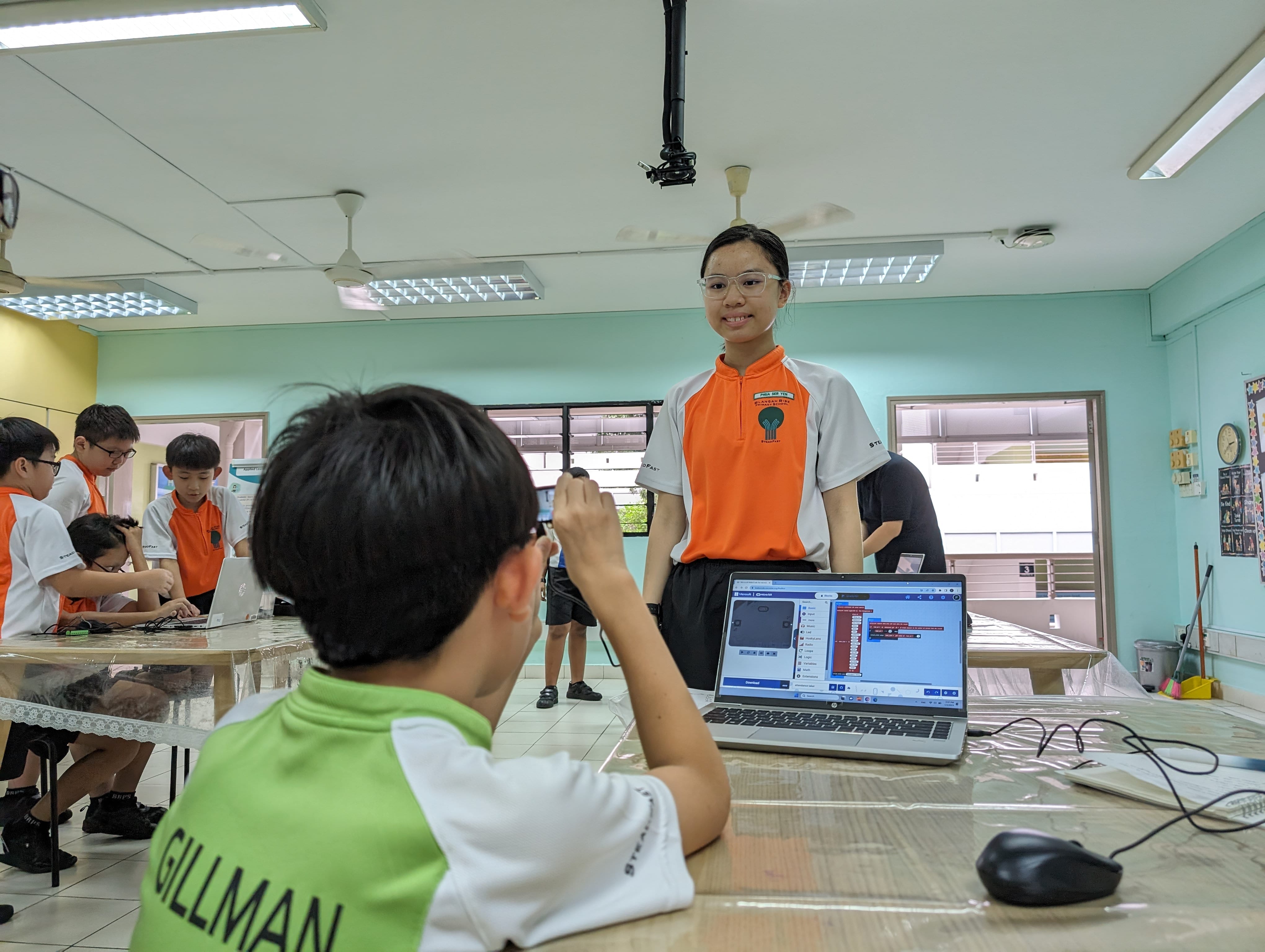 Students in school uniforms work on laptops in a classroom, with a girl in orange smiling at the camera.