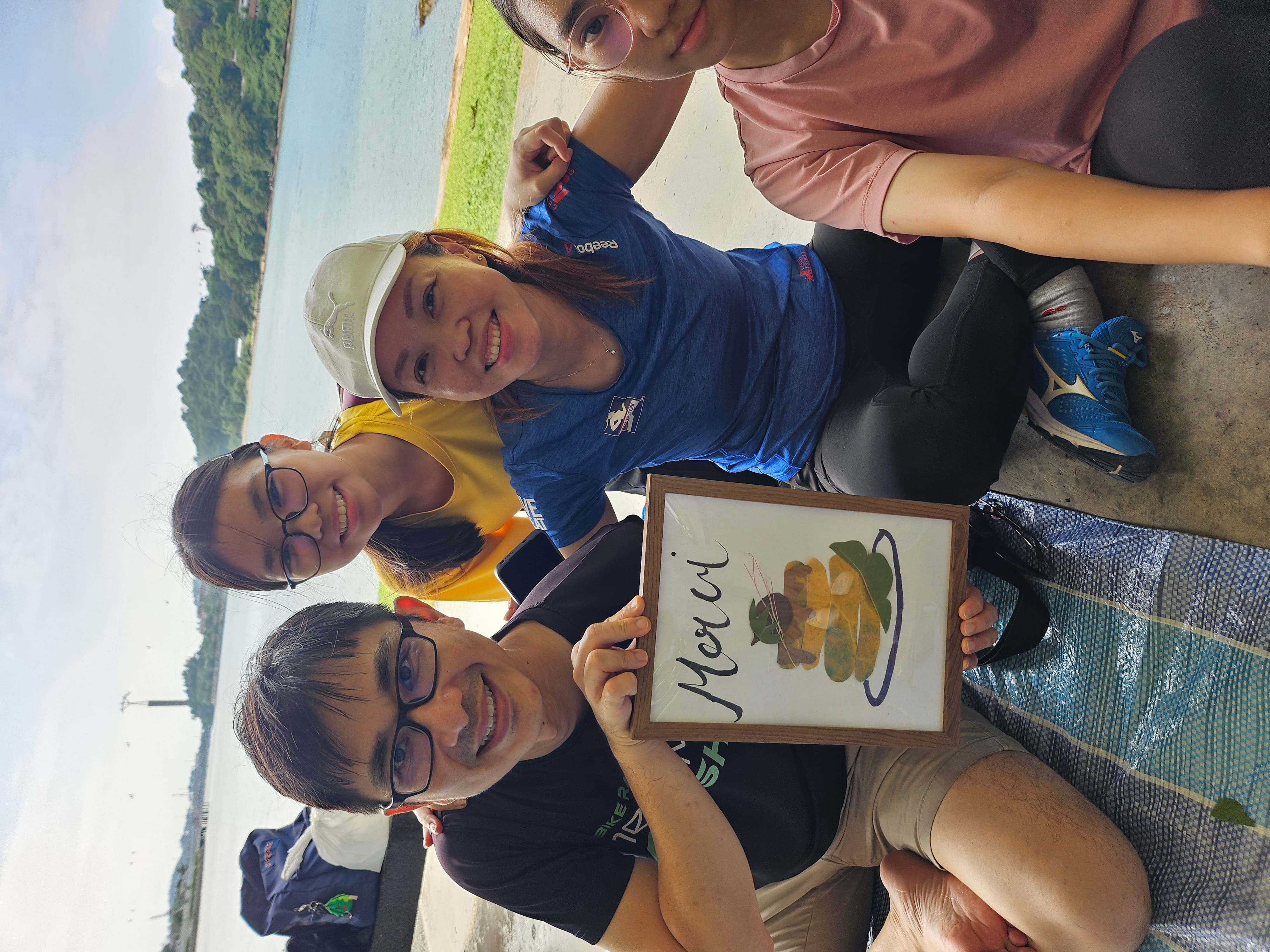 Four smiling people sit outdoors near a river, one holding a framed card that reads "Merci" with a food illustration.