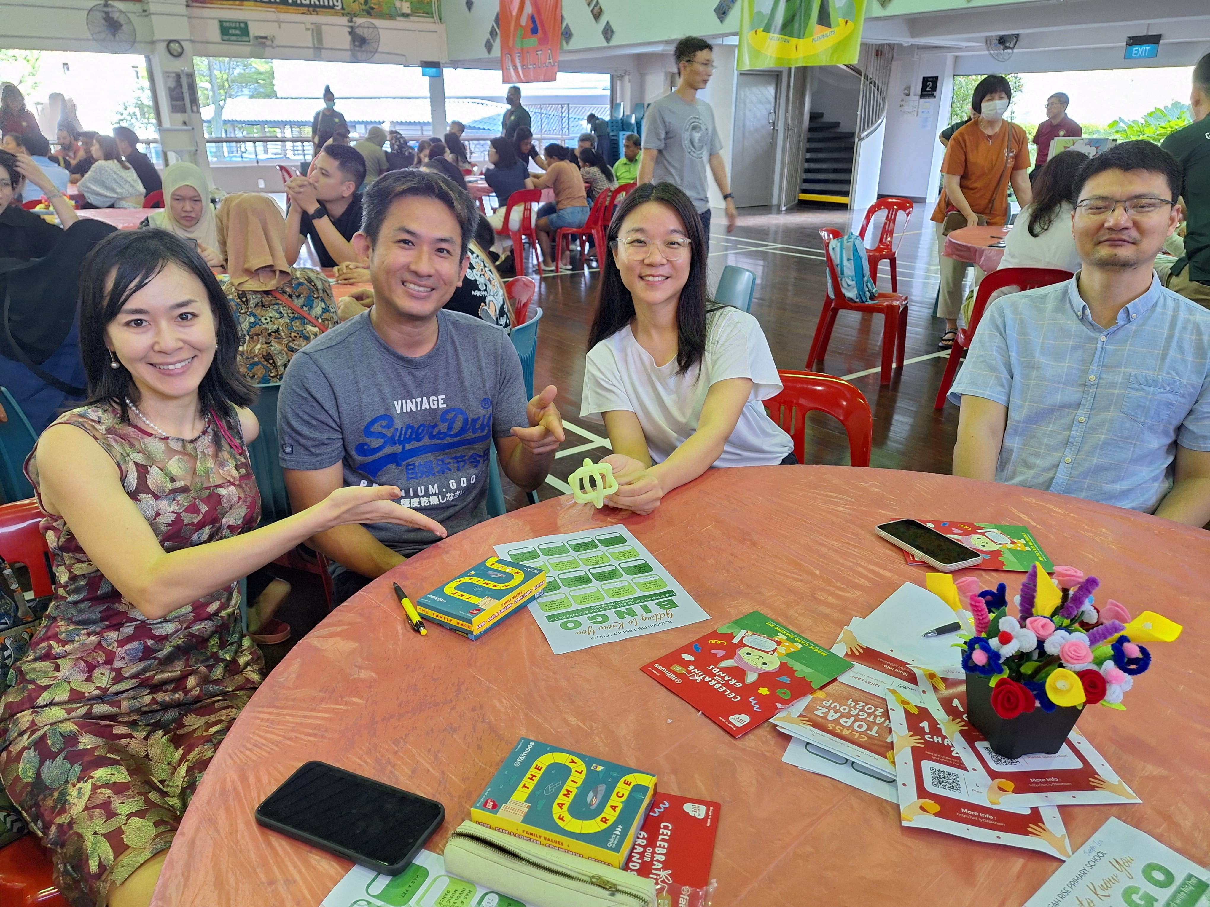 Four people smile at a round table with crafts and books in a busy community hall with red chairs.