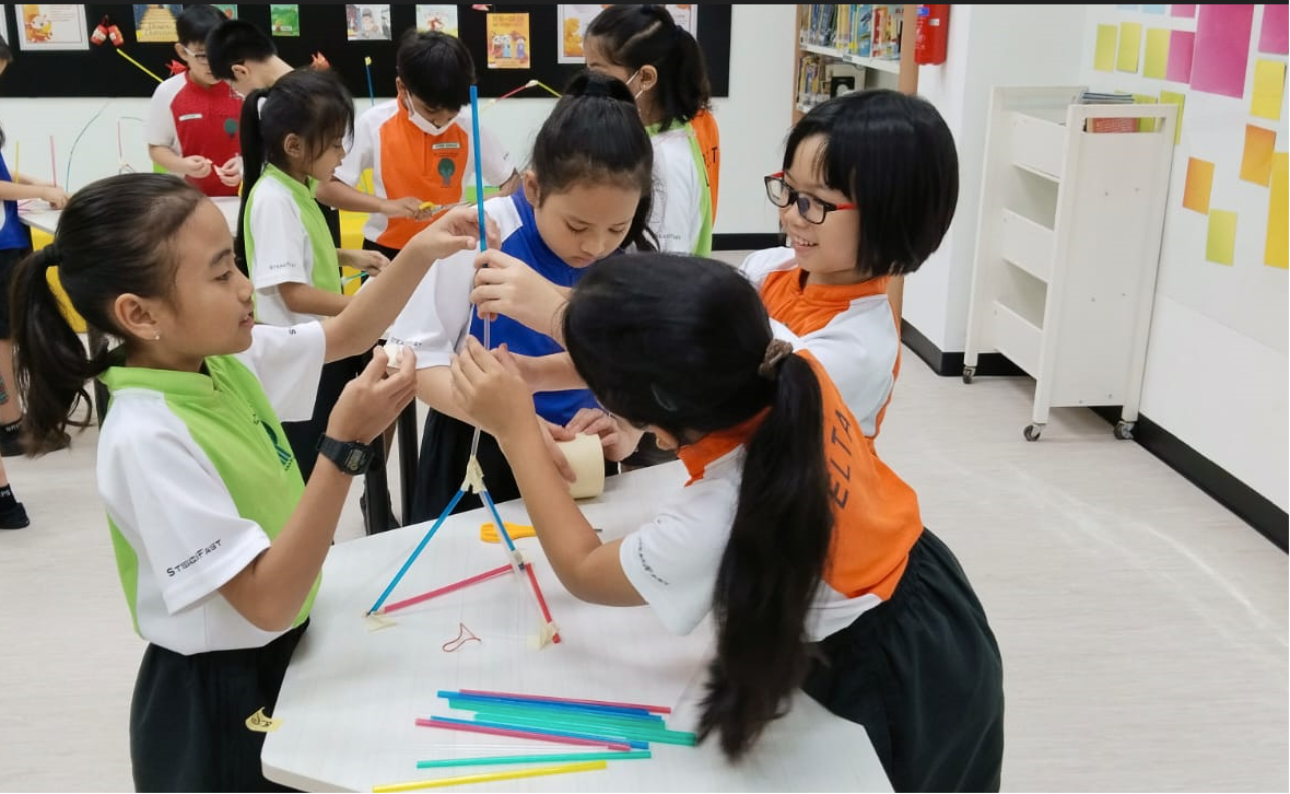 Children in school uniforms work together at a table, building a structure using colorful straws in a classroom.