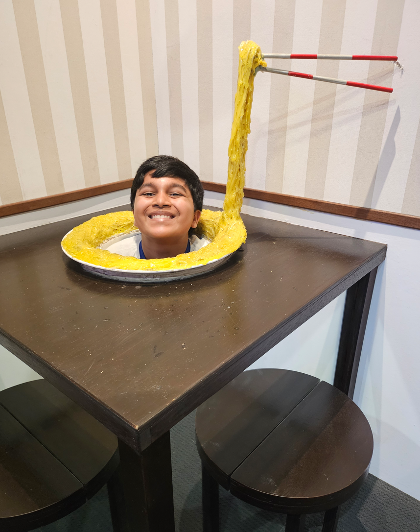 A smiling boy poses with his head through a table cutout designed to look like a noodle bowl with chopsticks.