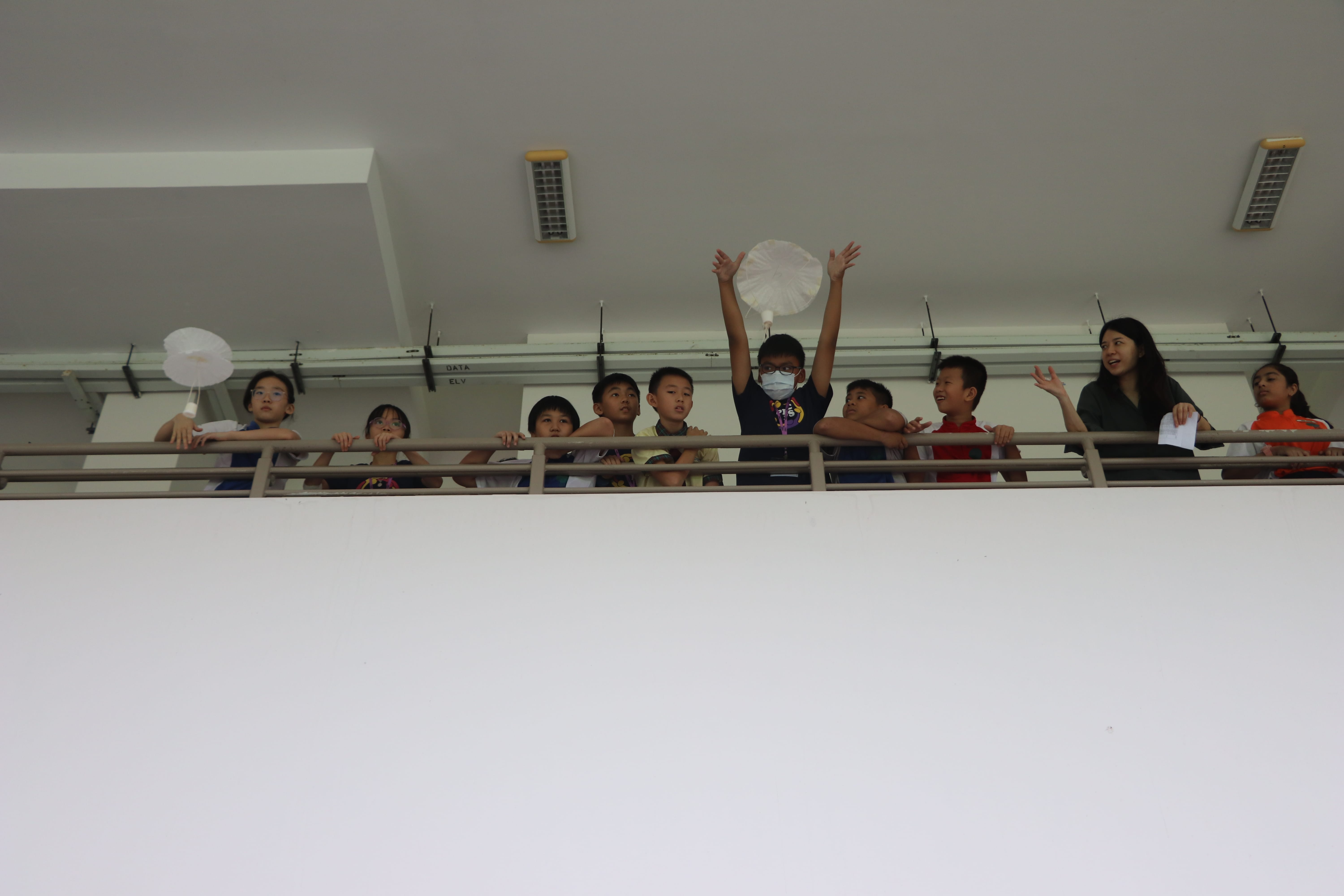 Children lean over a balcony railing; one raises both arms holding a white paper fan while an adult stands nearby.