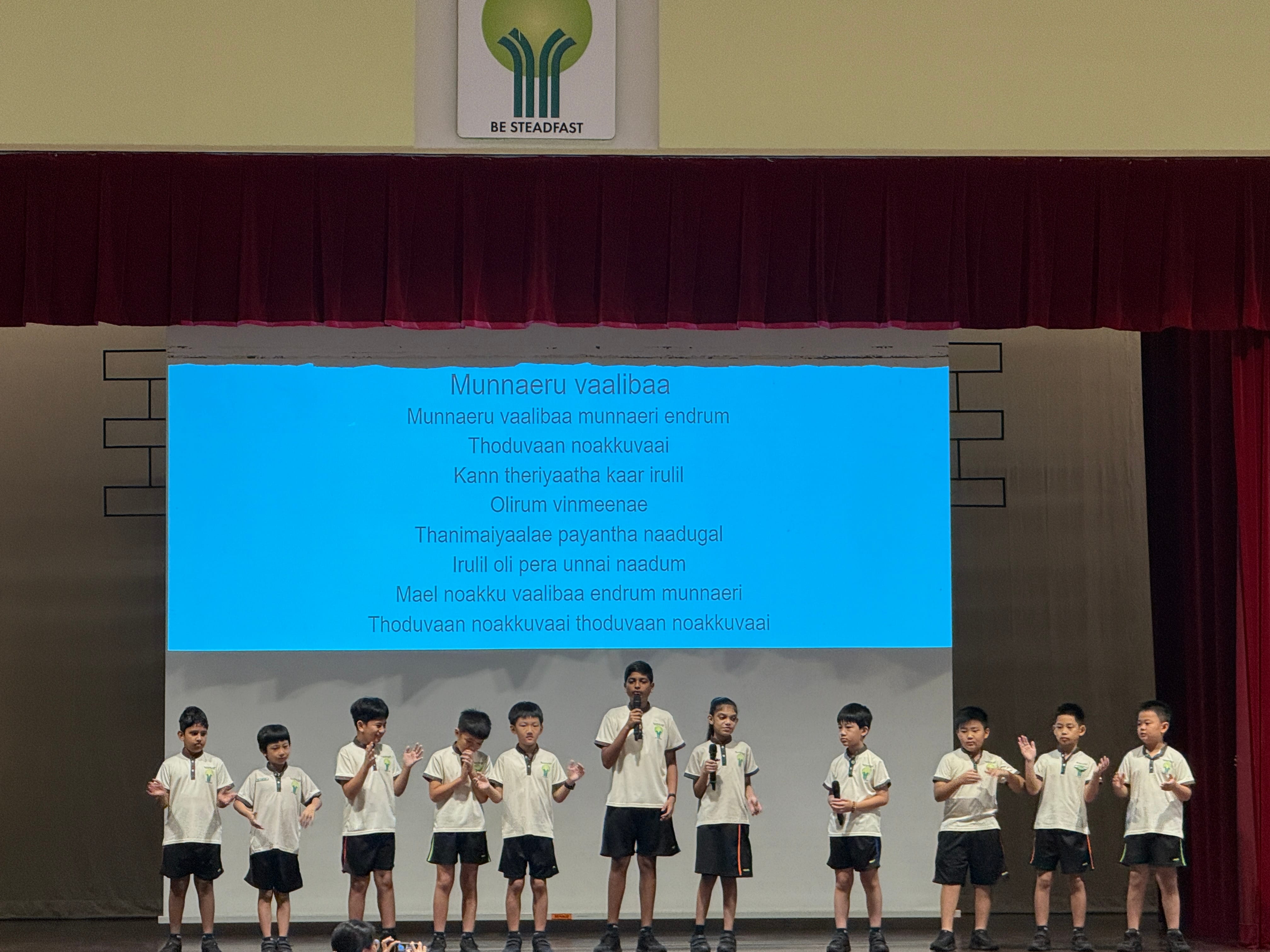 Ten boys in matching uniforms perform on a school stage with Tamil song lyrics projected on a blue screen behind them.
