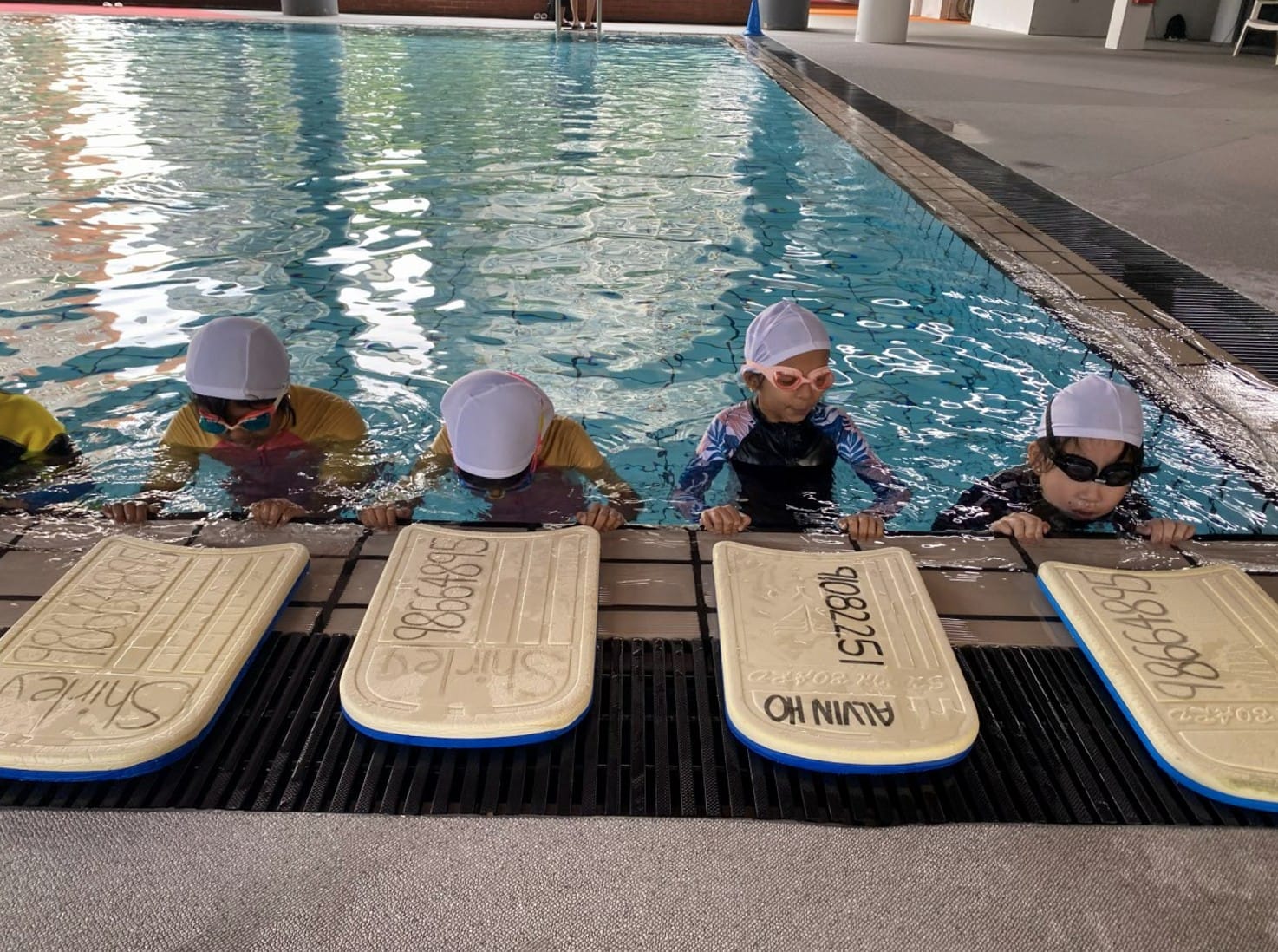 Four young children in swim caps and goggles hold kickboards at the edge of an indoor pool.