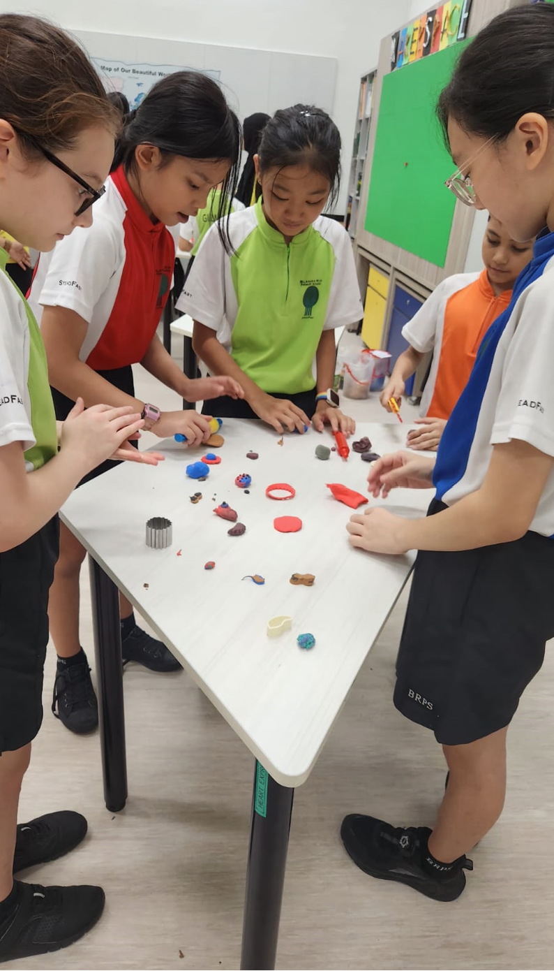 Four school children in uniforms stand around a white table molding colorful clay shapes in a classroom.