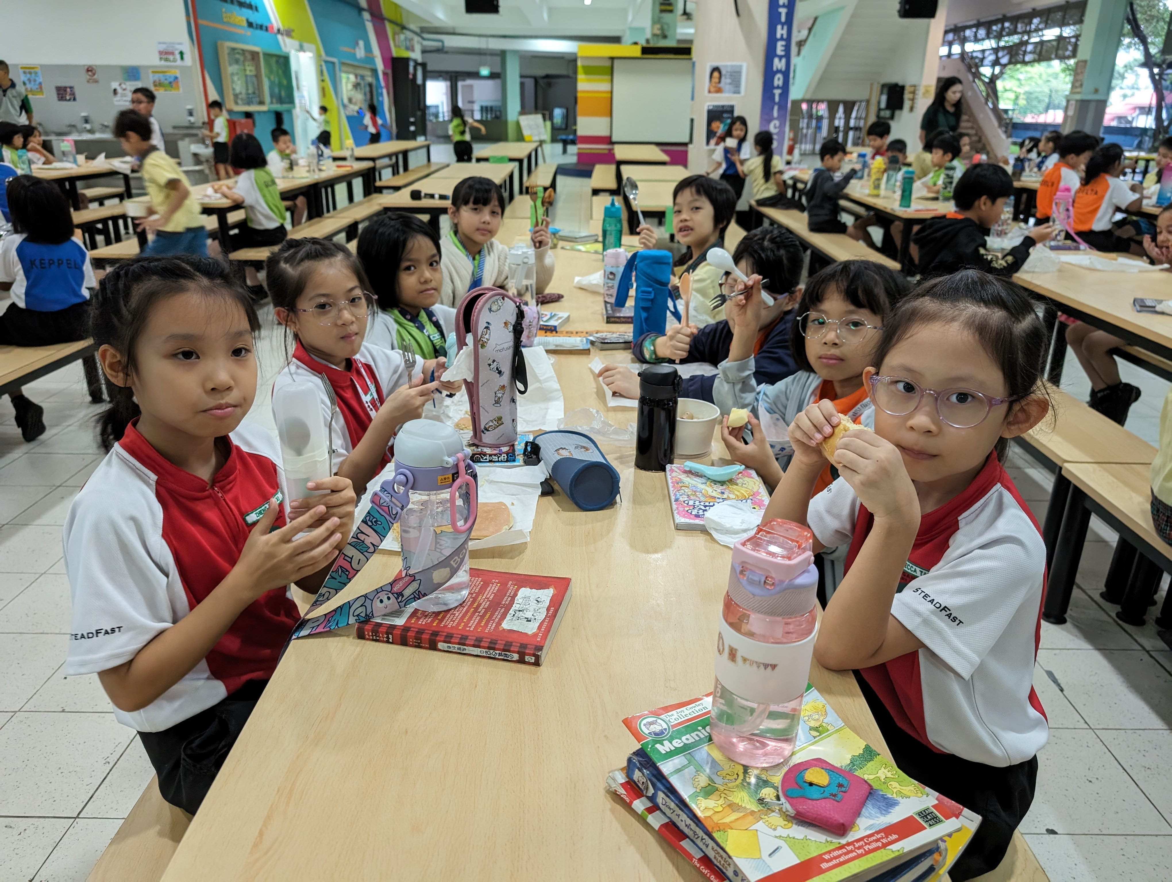 Young students in school uniforms eating lunch at long tables in a busy school cafeteria with books and water bottles.