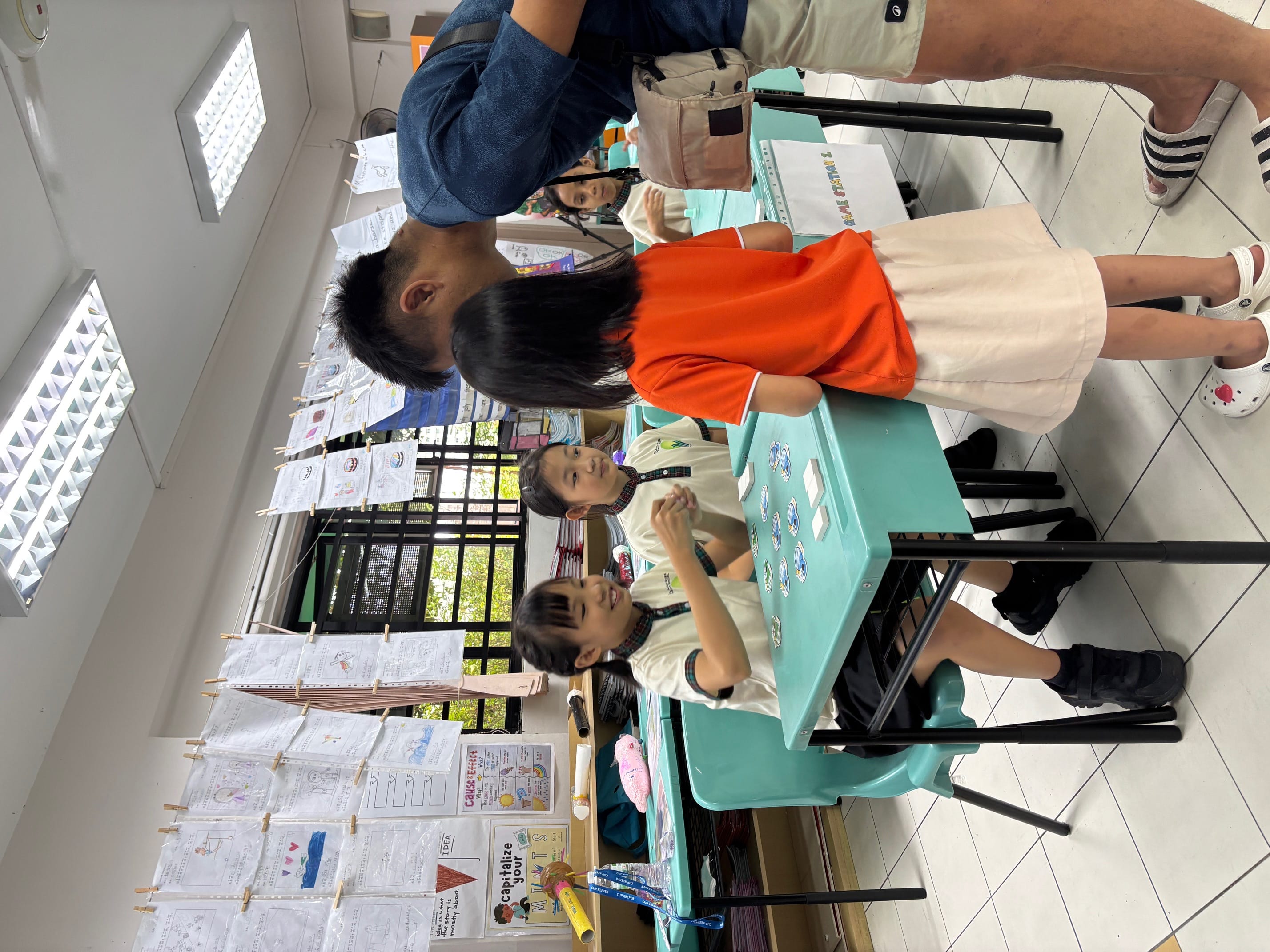 Children in school uniforms play a card game at a teal desk while an adult leans over to watch in a decorated classroom.