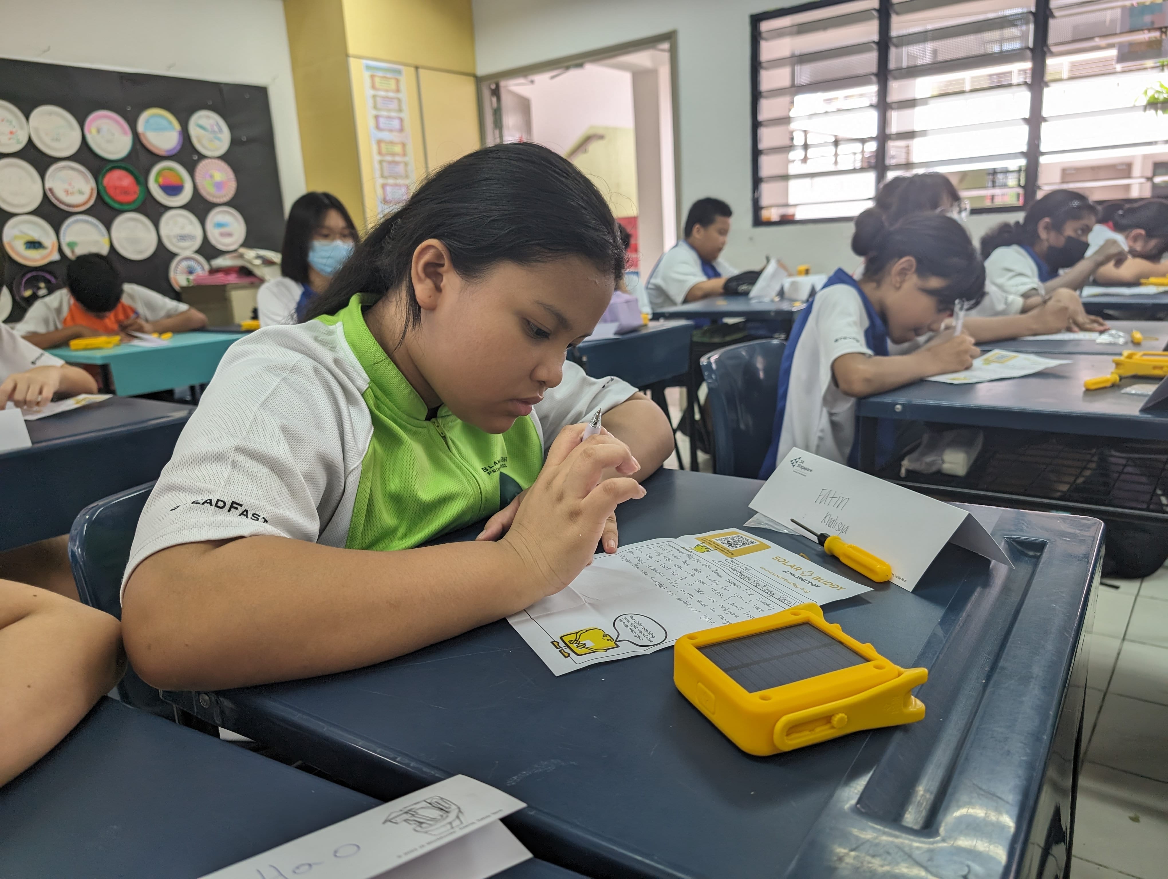 A girl in a green shirt writes at a school desk with a yellow Solar Buddy kit beside her, classmates working in background.