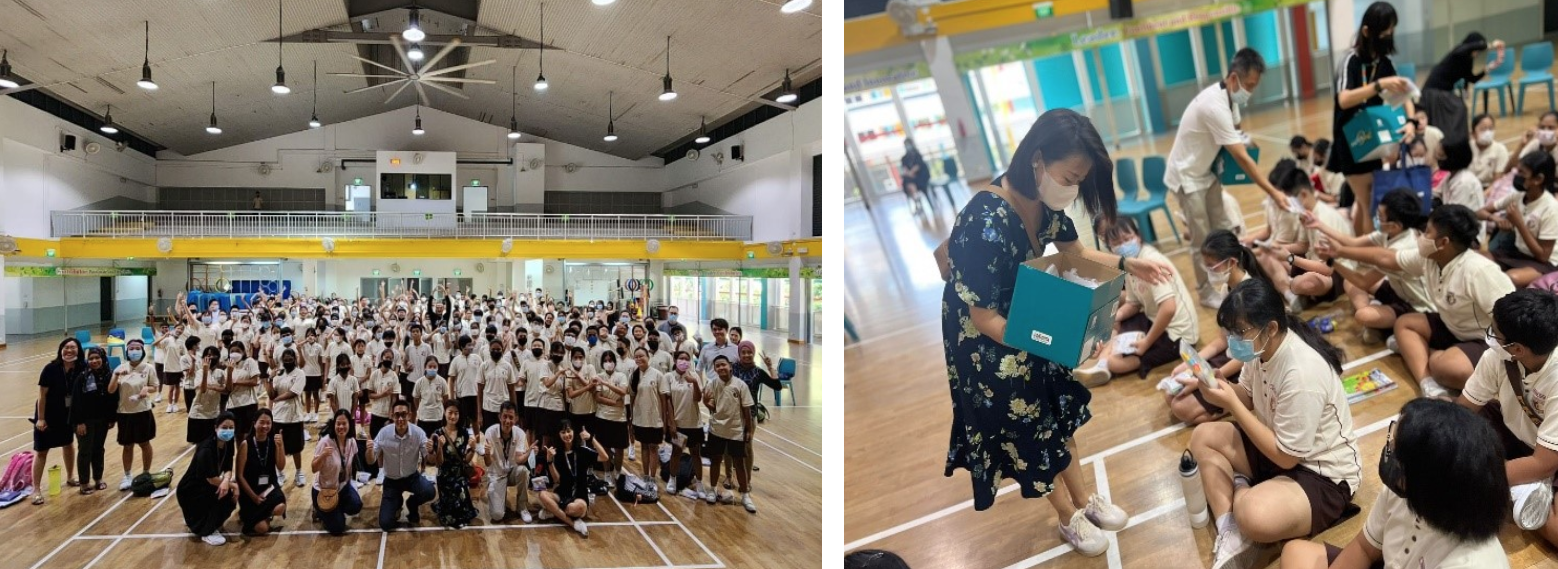 Large group of masked students in uniform pose in a gym; a woman distributes items from a box to seated students.