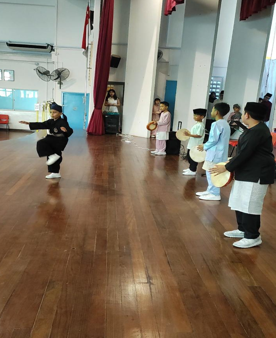 A student performs a traditional dance pose in black costume, others holding hand drums nearby in a hall.