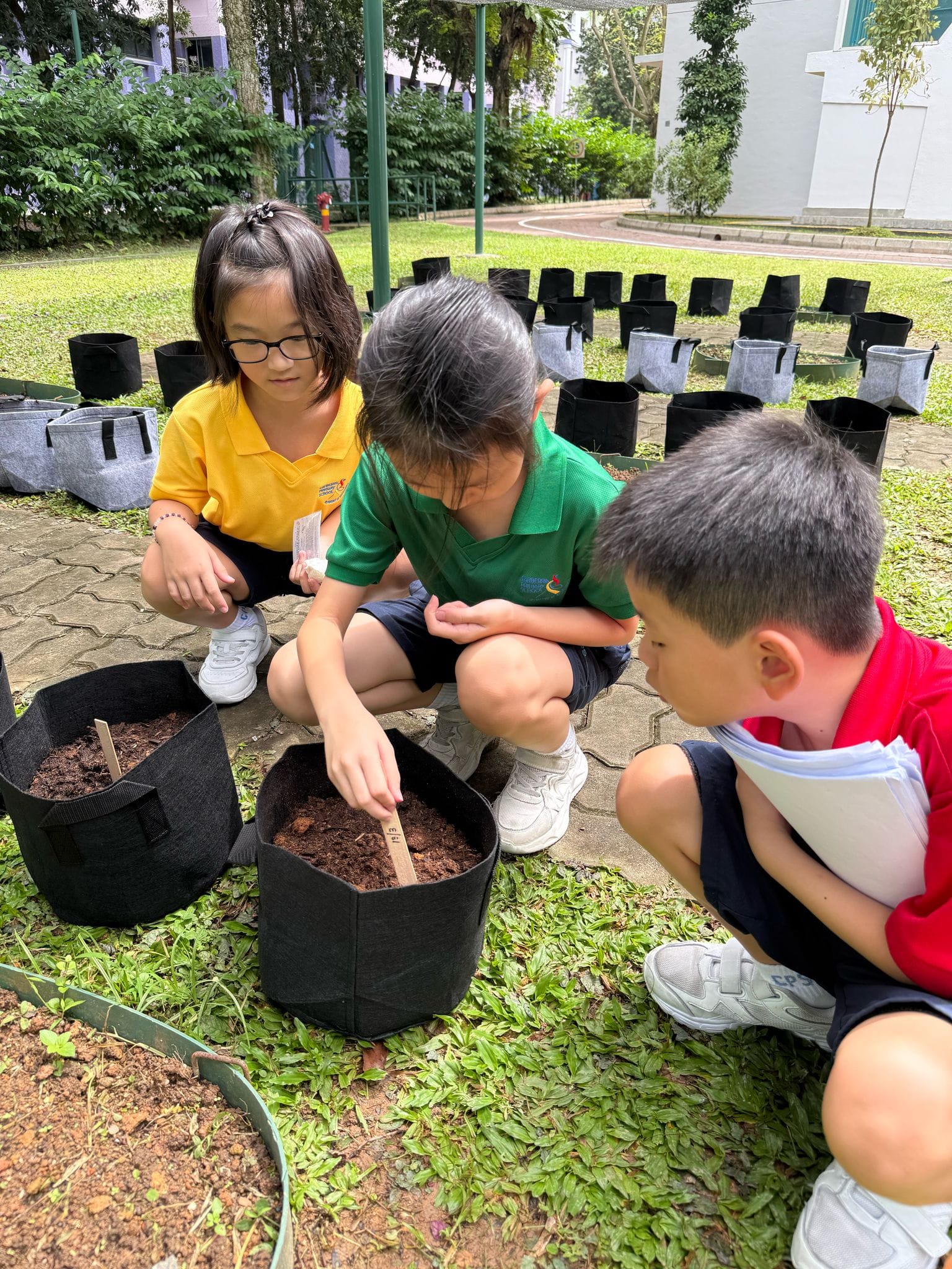 Three young children crouch outdoors around a black fabric pot filled with soil, placing a small label stick into it.