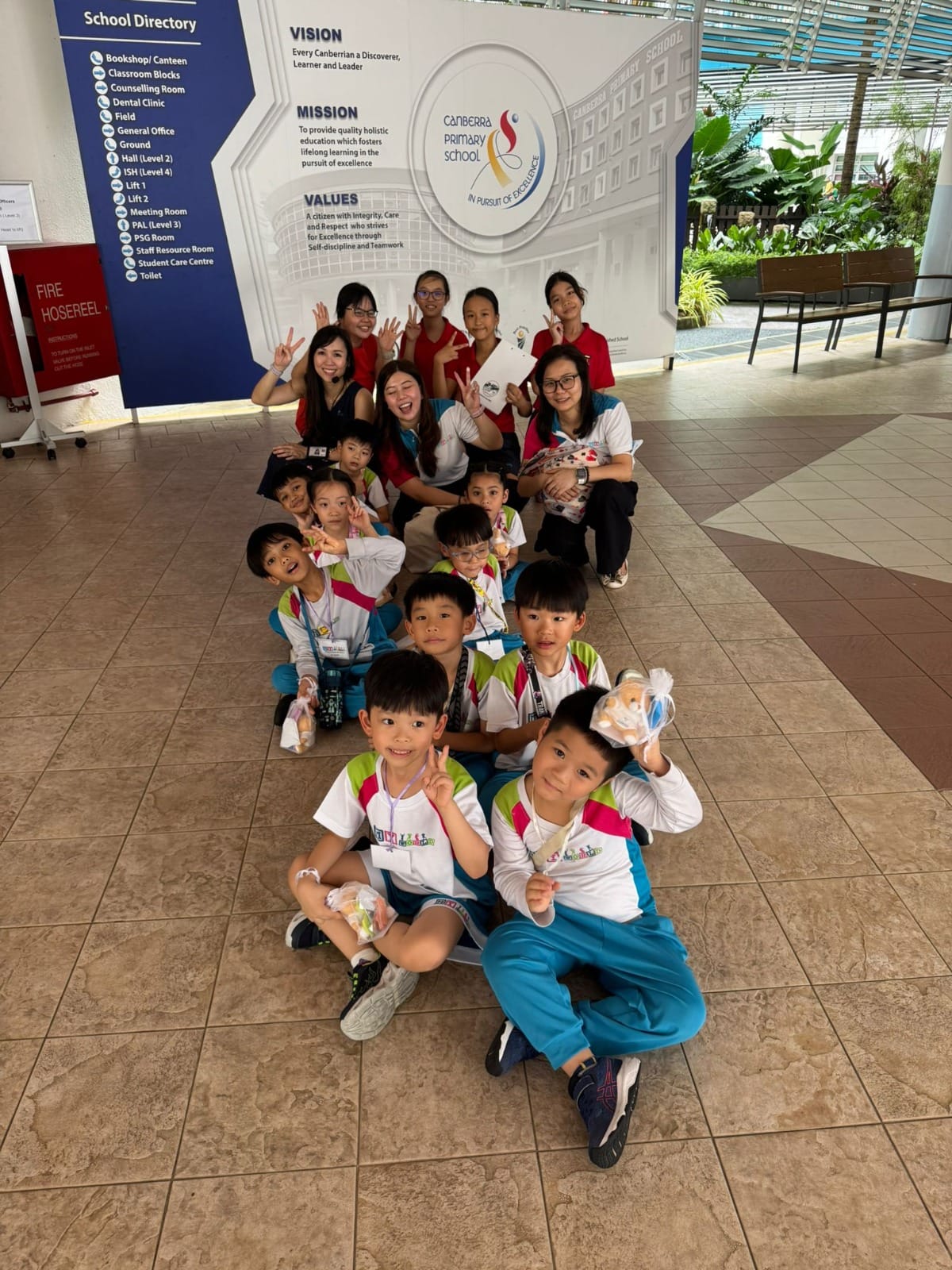 Students and teachers pose together on the floor before a 'Canberra Primary School' vision and mission board.