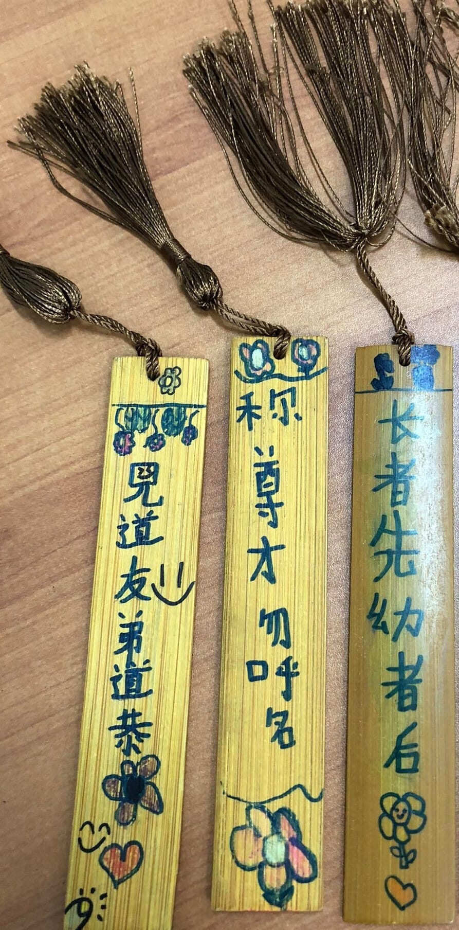 Three wooden bookmarks with Chinese characters, hand-drawn flowers, and gold tassels on a wooden surface.