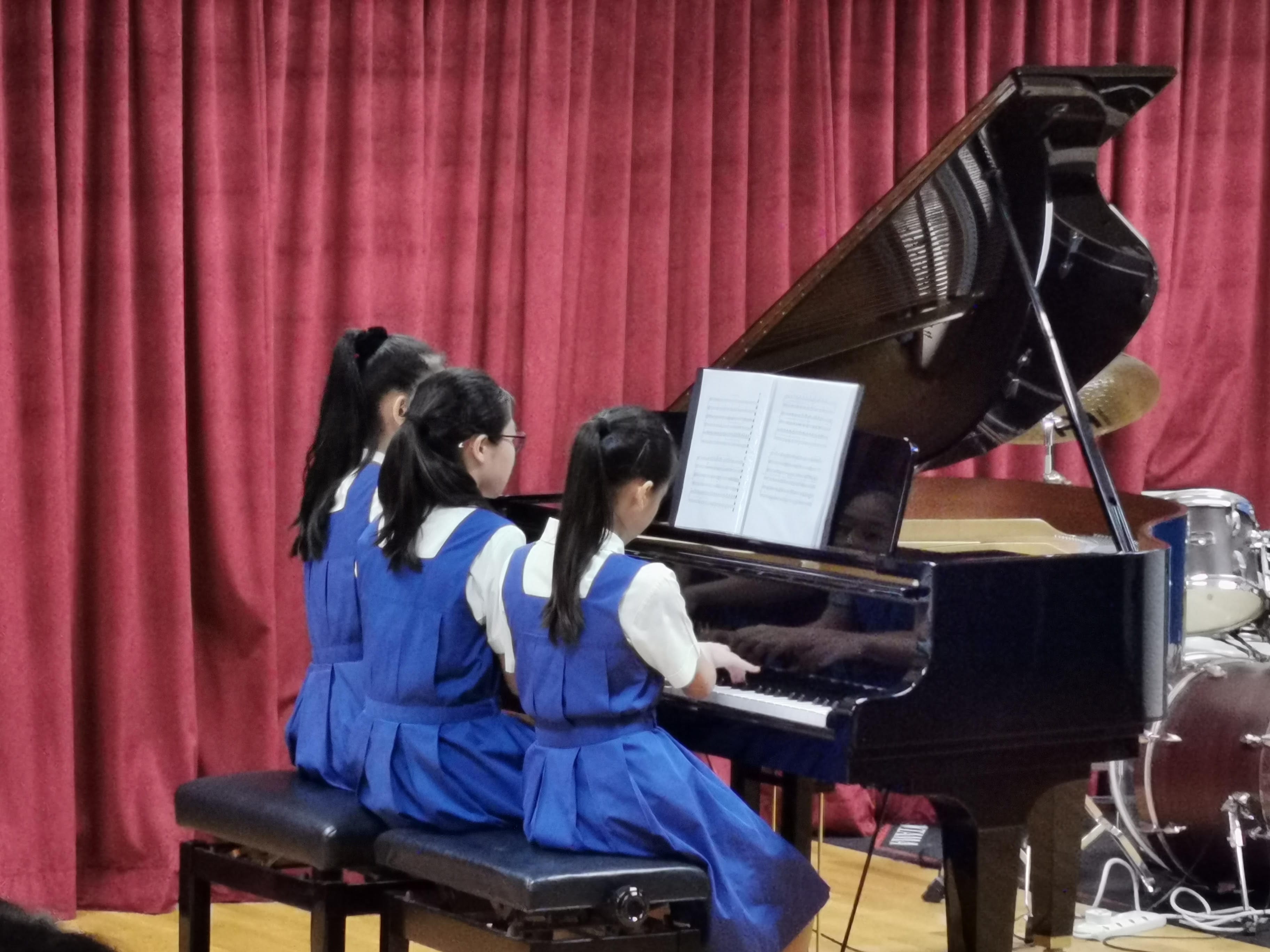 Three girls in blue uniforms sit together playing a grand piano on stage before a red curtain.