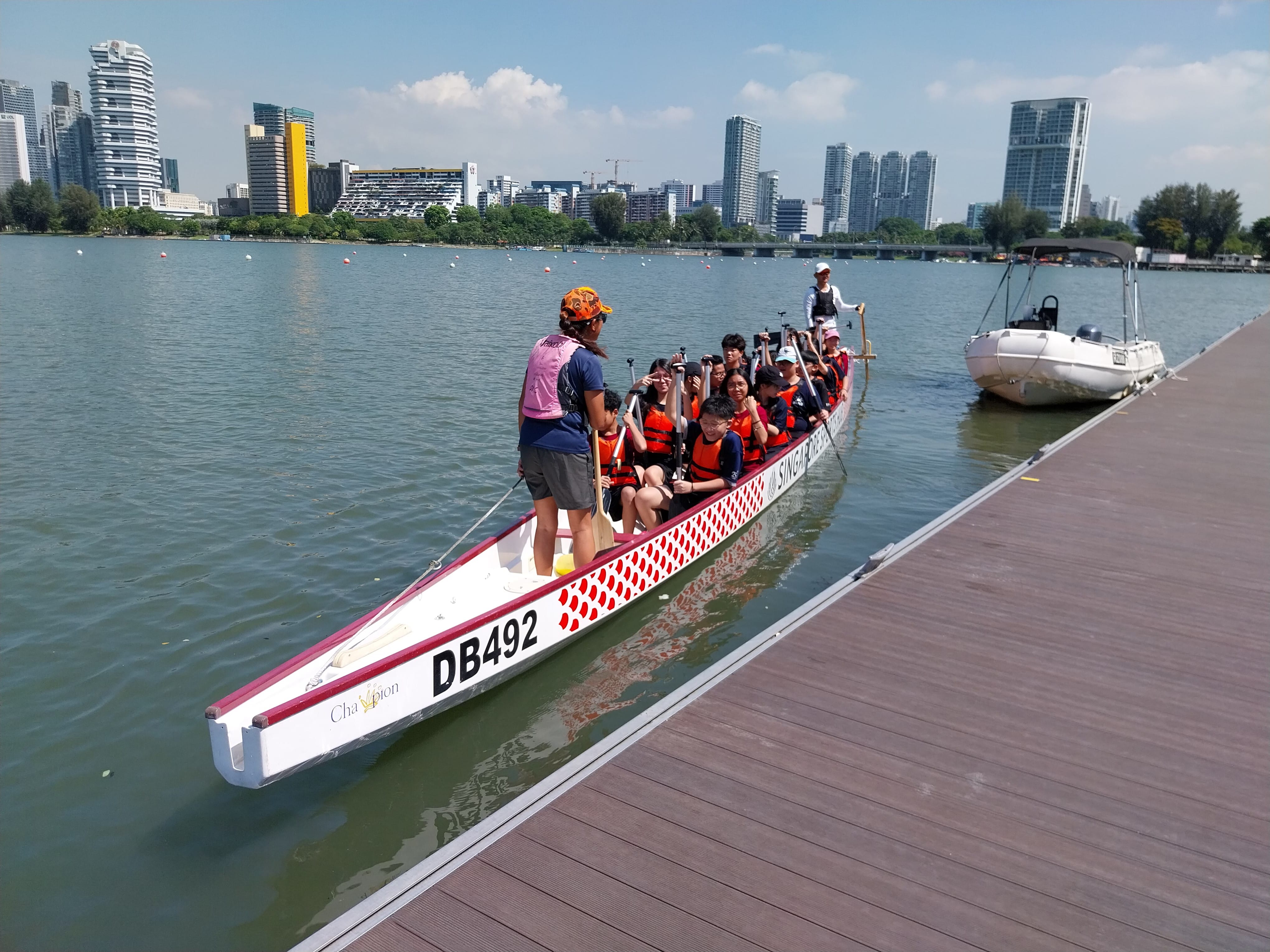 A group wearing orange life jackets sits in dragon boat DB492 beside a dock, with a city skyline in the background.